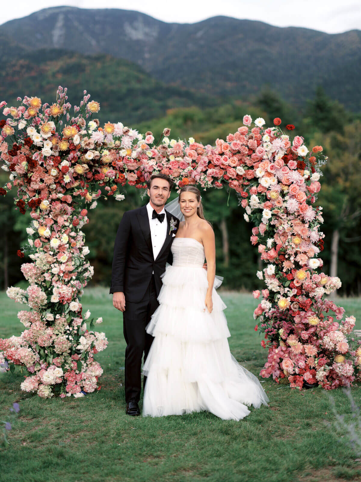 The bride and the groom are smiling, with an arch full of flowers and mountains in the background at The Ausable Club, NY.