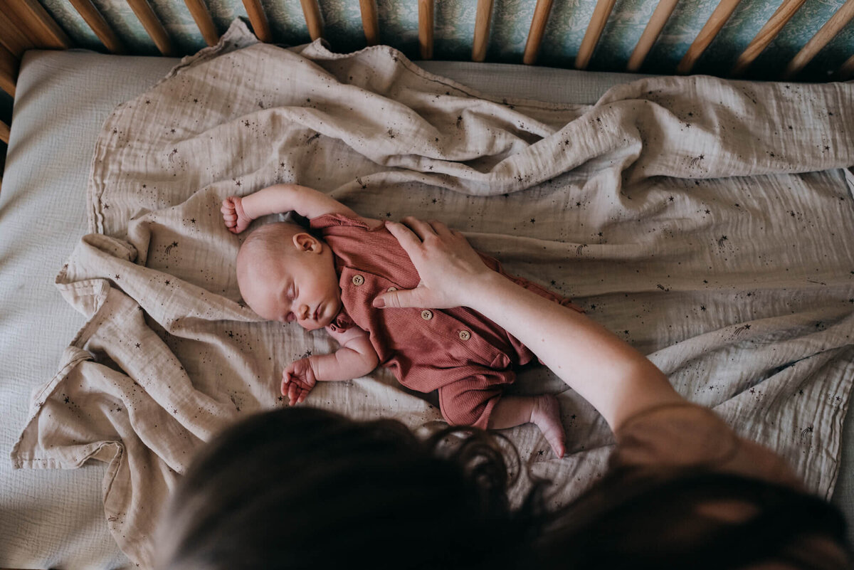 A mother helps position her sleeping newborn daughter in her crib during her photoshoot with Boston Newborn Photographer, Allison Wolf