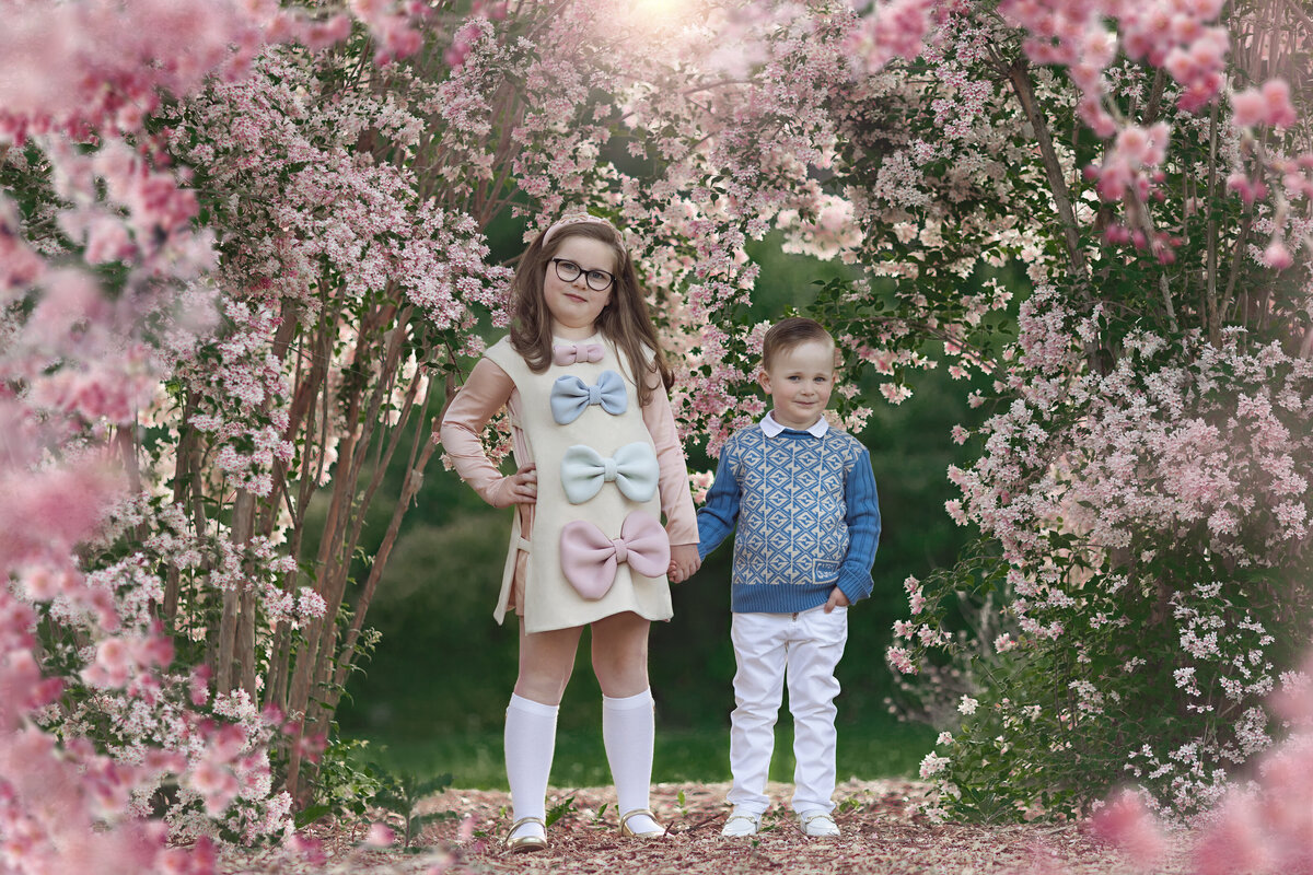 A toddler girl in a dress with giant bows holds hands with her toddler brother in a park surrounded by pink flowers
