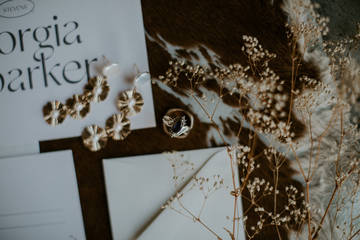 A pair of earrings and wedding bands on white stationery with black font atop cow hide with dried flower stems.