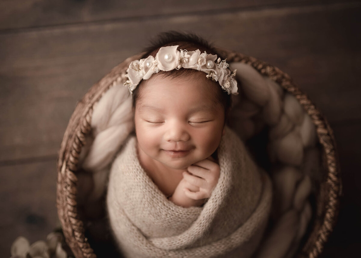 smiling newborn baby girl in dark rattan basket with beige filler and wrap with beautiful soft light falling across her smiling face