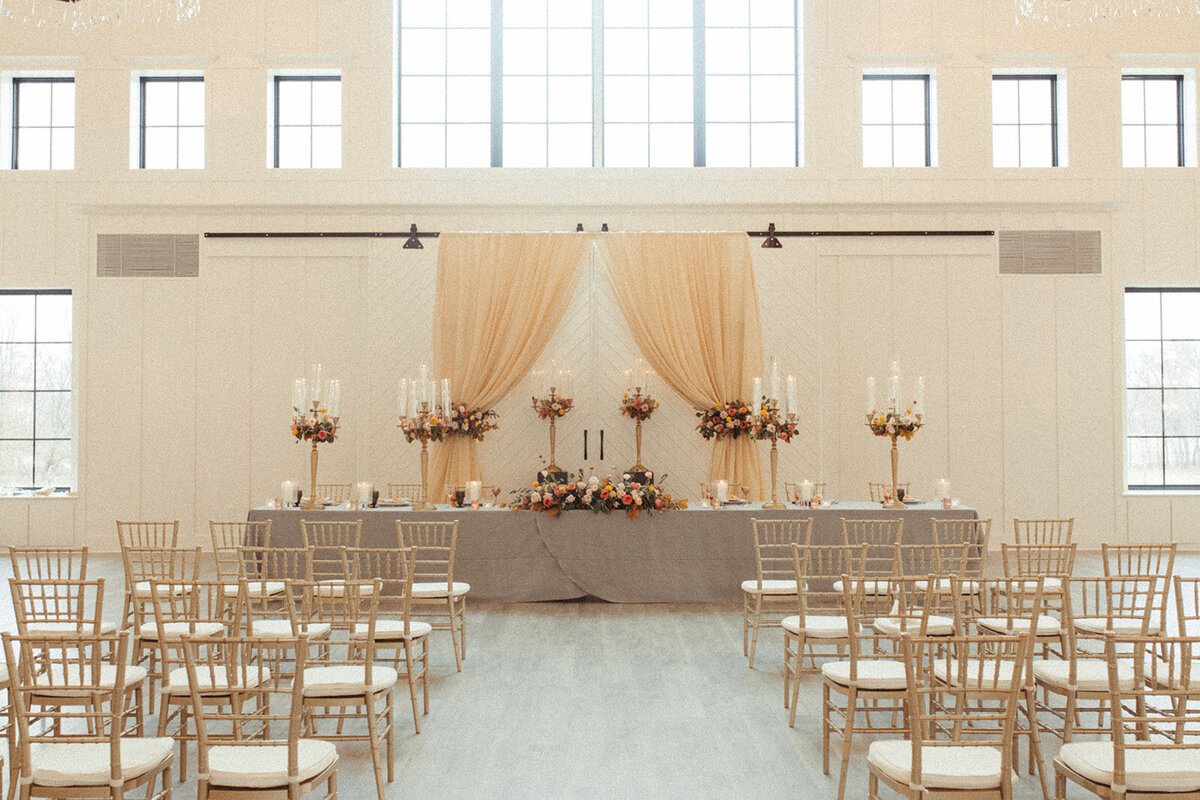 A bright and airy wedding reception room filled with gold chairs, tall candlesticks and peach curtains on a wall.