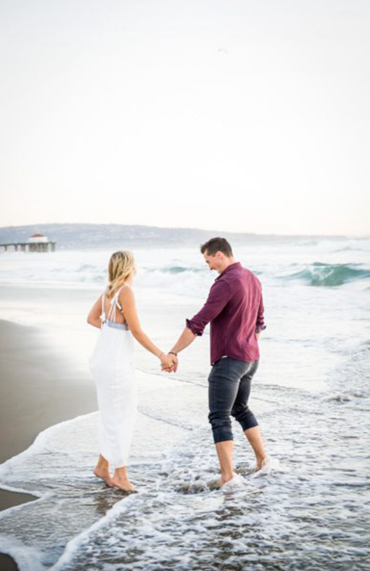 A man and woman face away from the camera, holding hands and playing in the ocean for their engagement session.