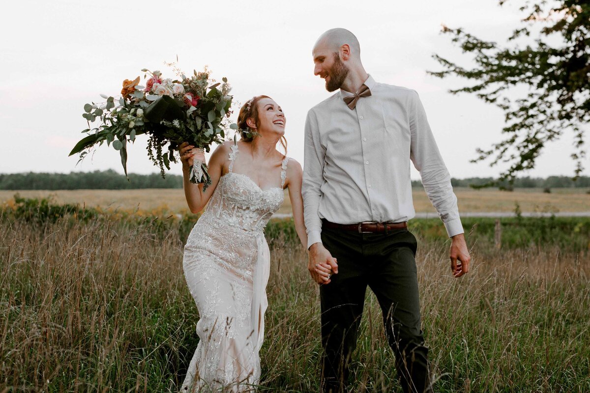 Bride and groom walk through Huron County fields looking at each other and holding hands for casual wedding day photo. The bride holds a large bouquet in the other hand.