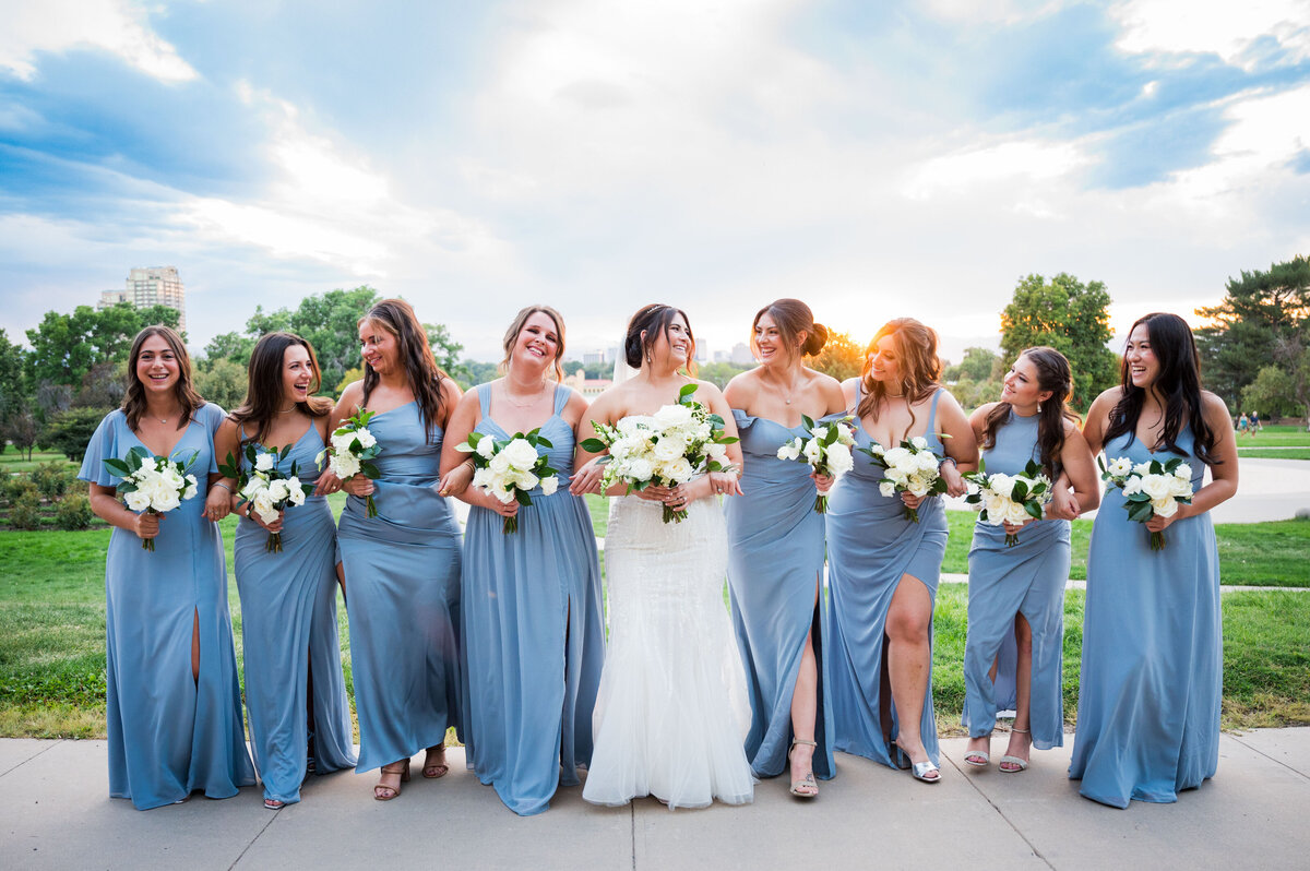 A bride links arms with her bridesmaids who are wearing blue dresses, captured by Colorado wedding photographer, Two One Photography.