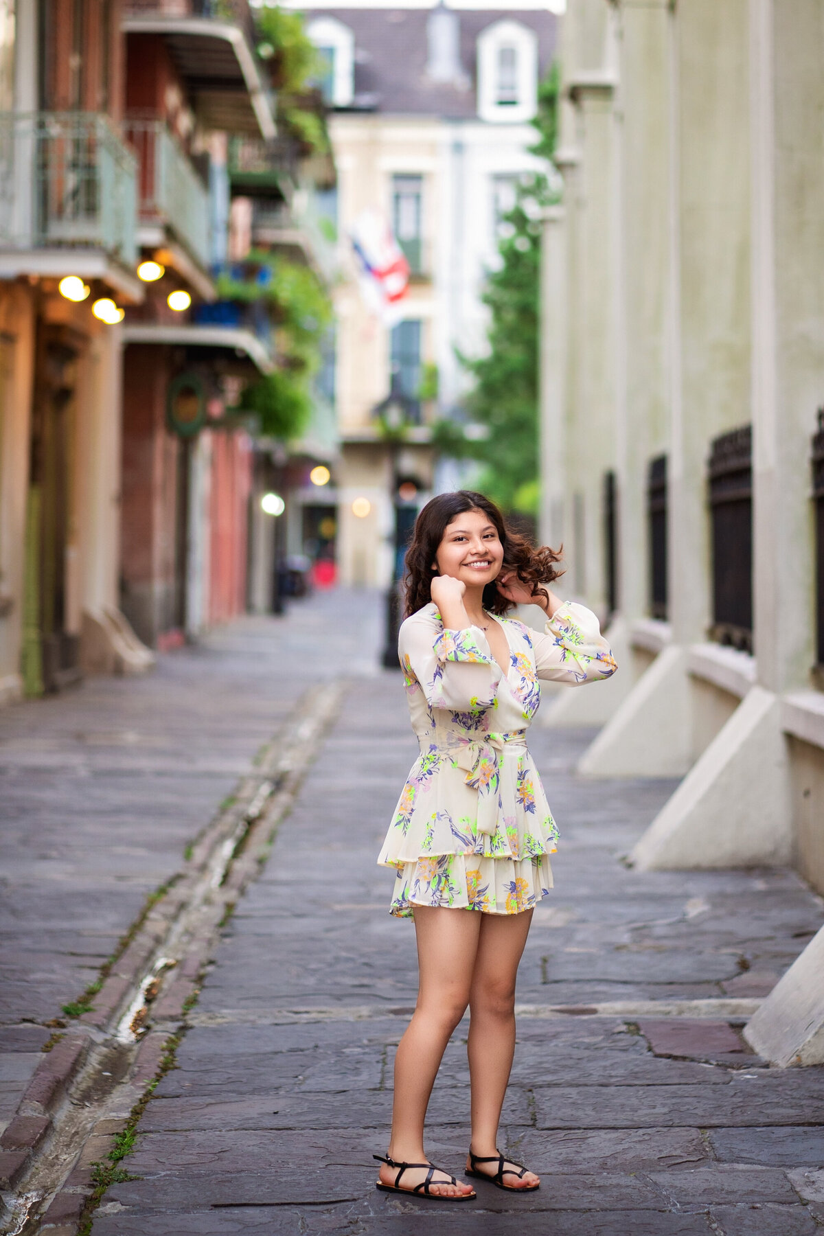 High School senior hispanic female in Pirate's Alley in New Orleans.  She is wearing a short jumper, sandals, and is swinging her hair and laughing.  The street is cobblestone and St. Louis Cathedral is on her right.