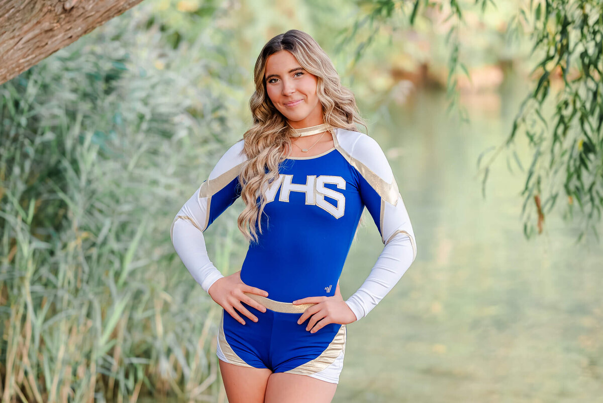A high school senior, wearing her blue, white and gold cheer uniform, puts her hands on her hips. She is standing in front of the lake at Oak Grove Lake Park in Chesapeake, VA.