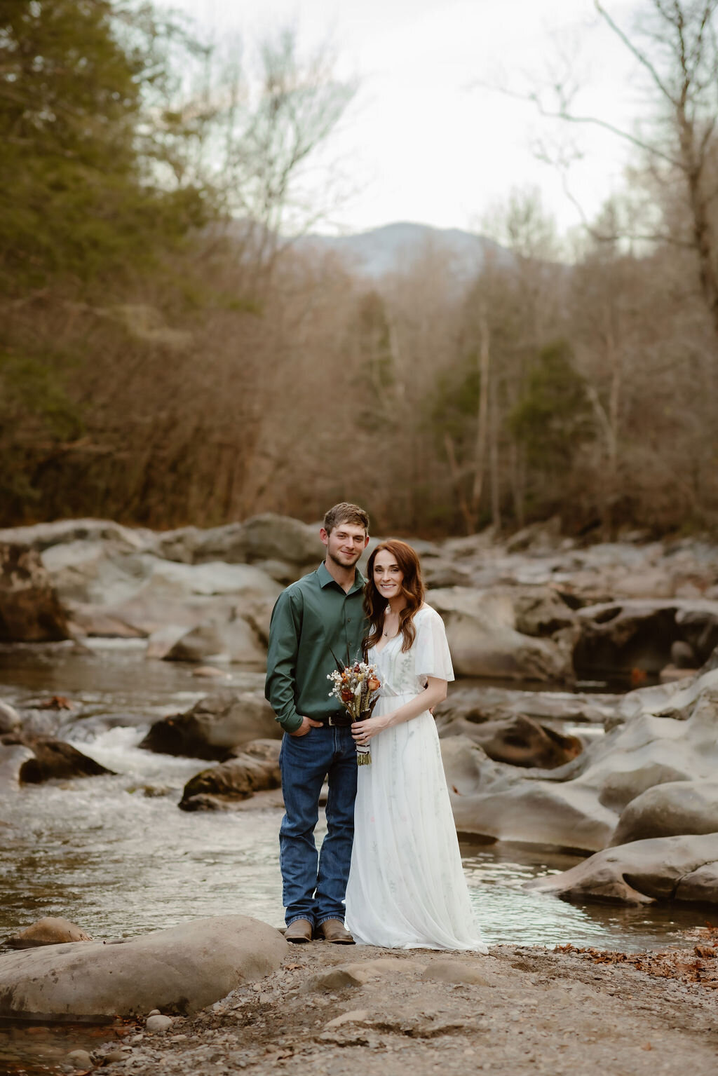 Bride and groom standing side by side on the riverbank at Greenbrier, holding a rustic bouquet with a mountain backdrop, during their eloping to Gatlinburg photo session.
