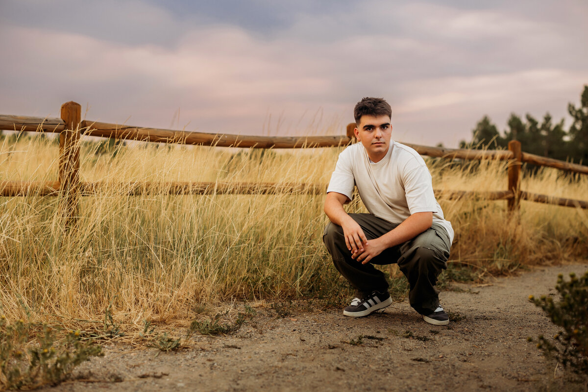 high school senior boy squats in the middle of a dirt path posing for his senior photos