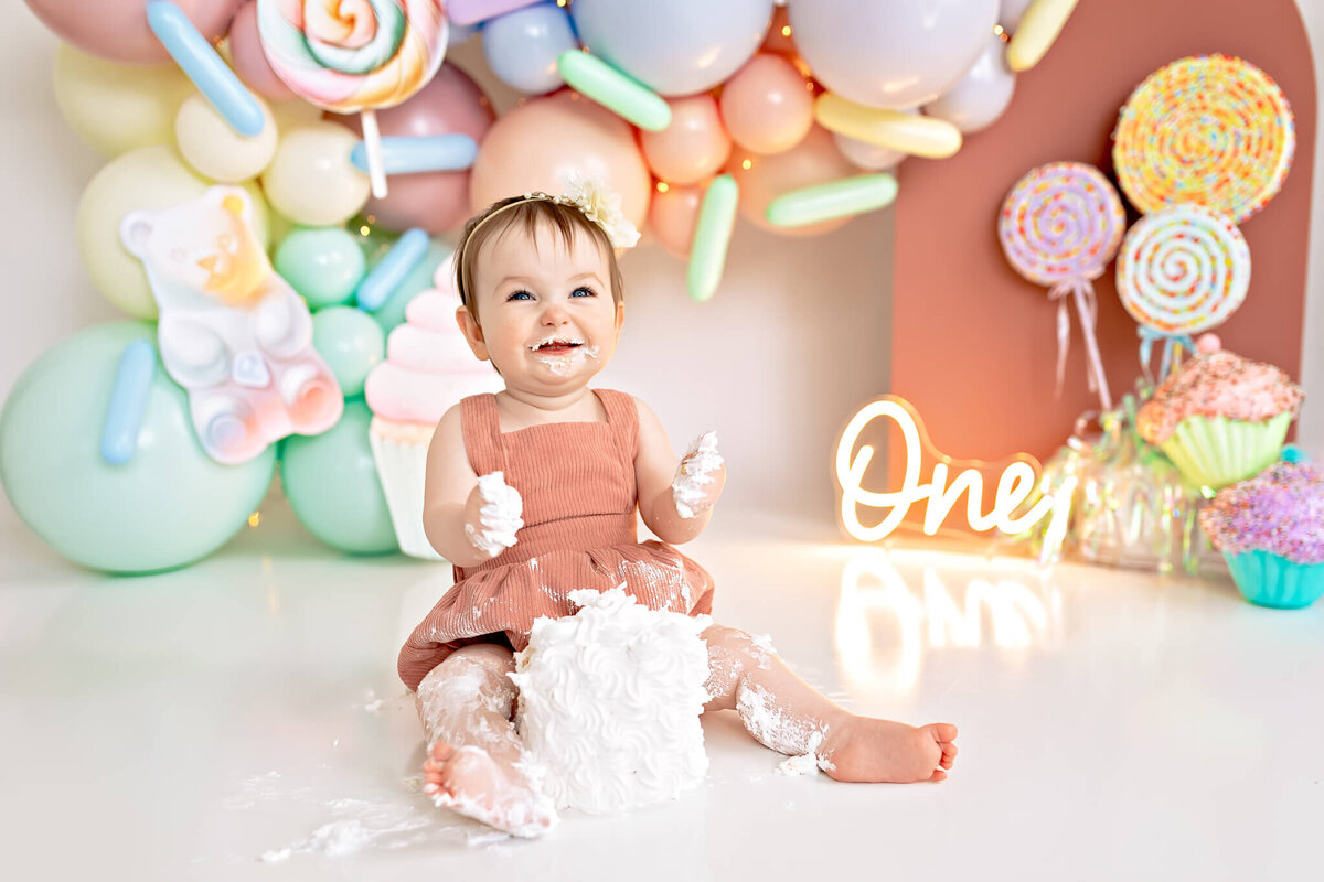 A smiling baby wearing a pink dress sits on the floor with a cake, surrounded by colorful balloon decorations and lollipops. A neon sign says "One." The setting is bright and playful, suggesting a first birthday celebration.