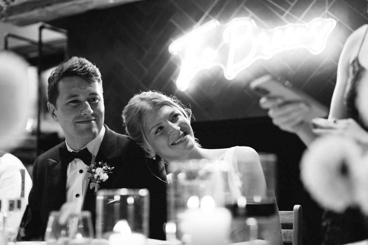 A couple, elegantly dressed, share a tender moment at an event as the woman rests her head on the man's shoulder. Nearby, a speaker engages the crowd while a neon "The Brews" sign glows in the background—captured perfectly by skilled wedding photographer in Muskoka, Jennifer van Son..