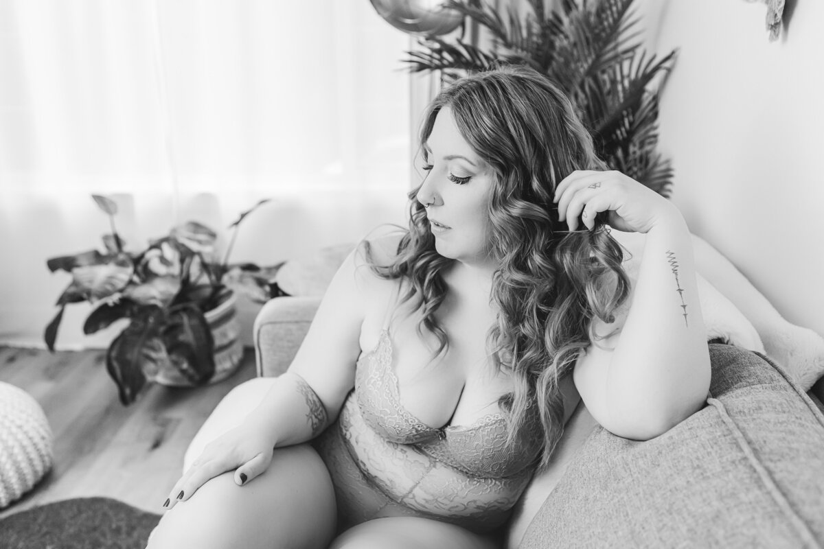 A woman sitting on a couch in light gray lingerie, gazing away in a reflective moment while twirling her hair.
