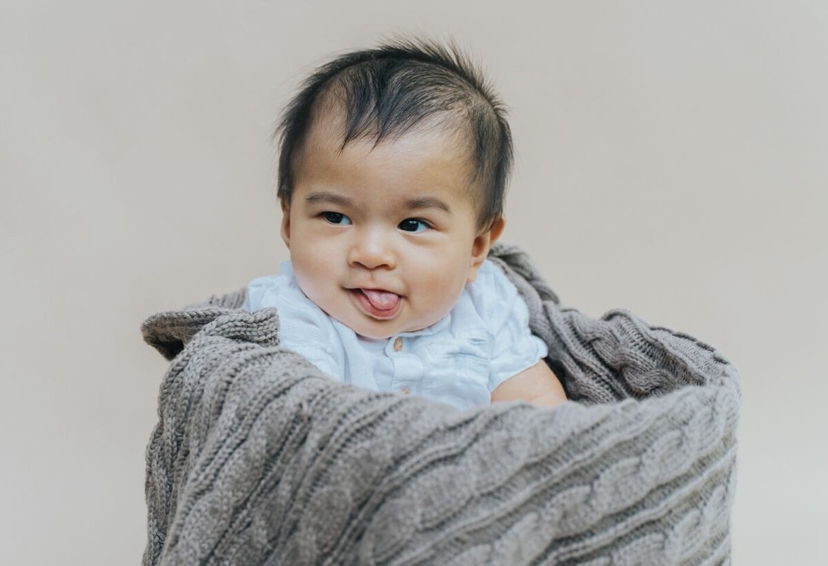 Baby boy wrapped in a chunky grey knitted blanket, wearing a light blue shirt and playfully sticking out his tongue.