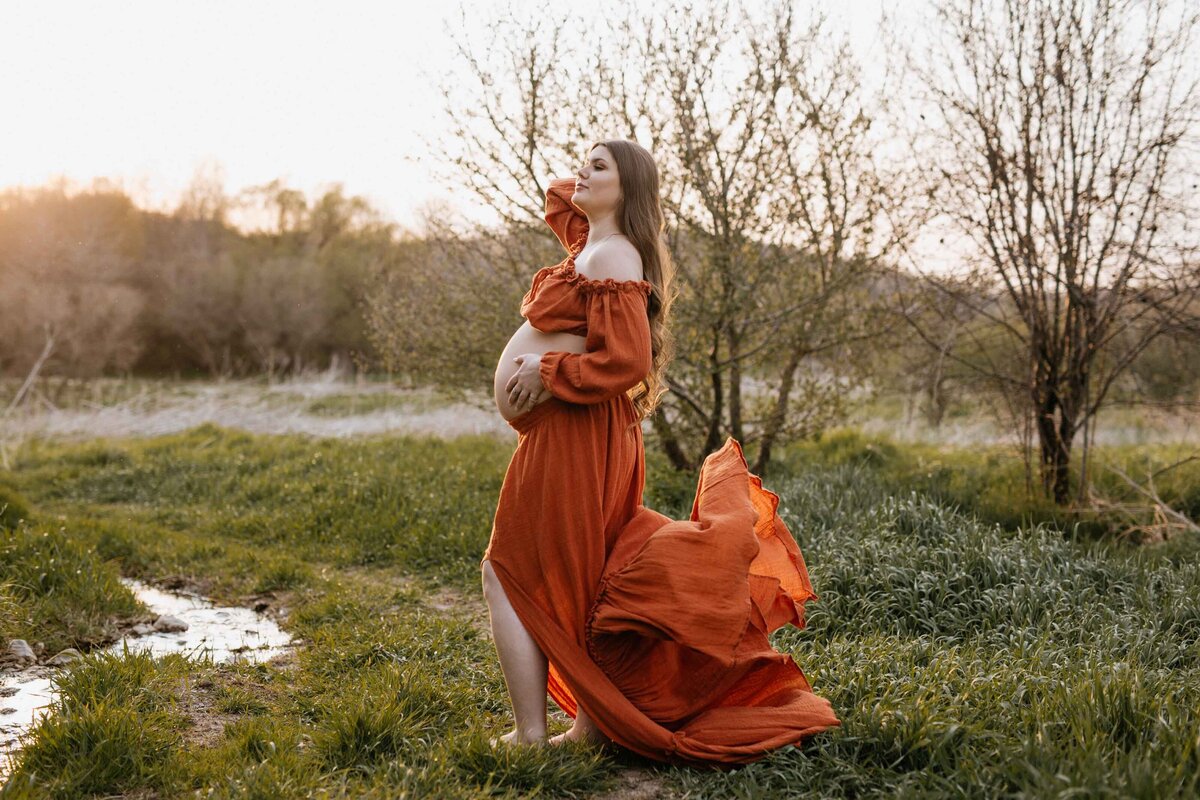 Expectant mom in London, Ontario outdoor maternity photoshoot. She is standing barefoot on a grassy trail in a two-piece maternity skirt and top, bump exposed. Her dress is blowing in the wind and she is pushing her hair off of her face.
