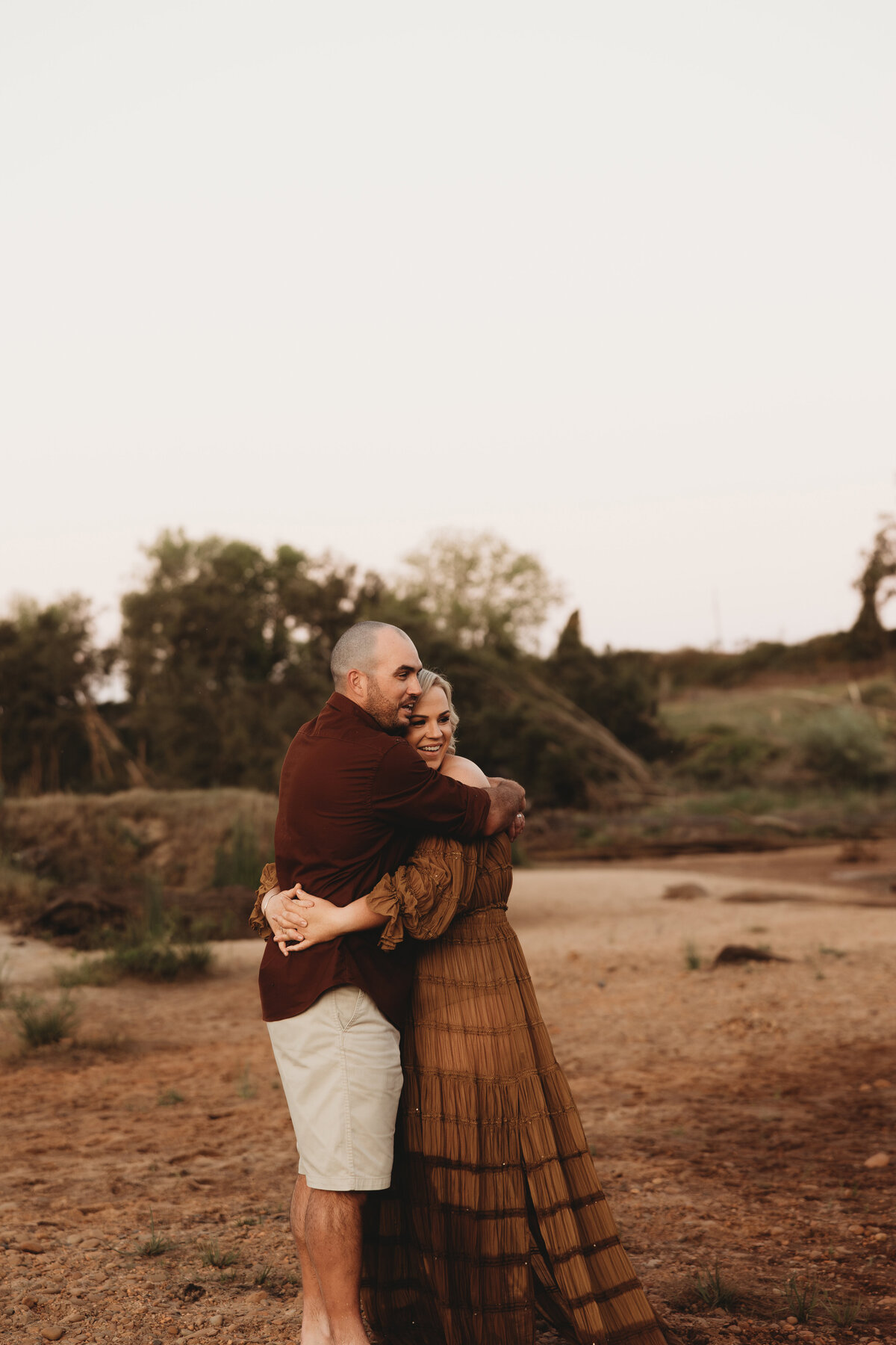 Two people stand outdoors on a dry, sandy surface, hugging each other. The backdrop includes patches of greenery and trees, offering the perfect setting for a heartwarming Wollongong maternity photography session.