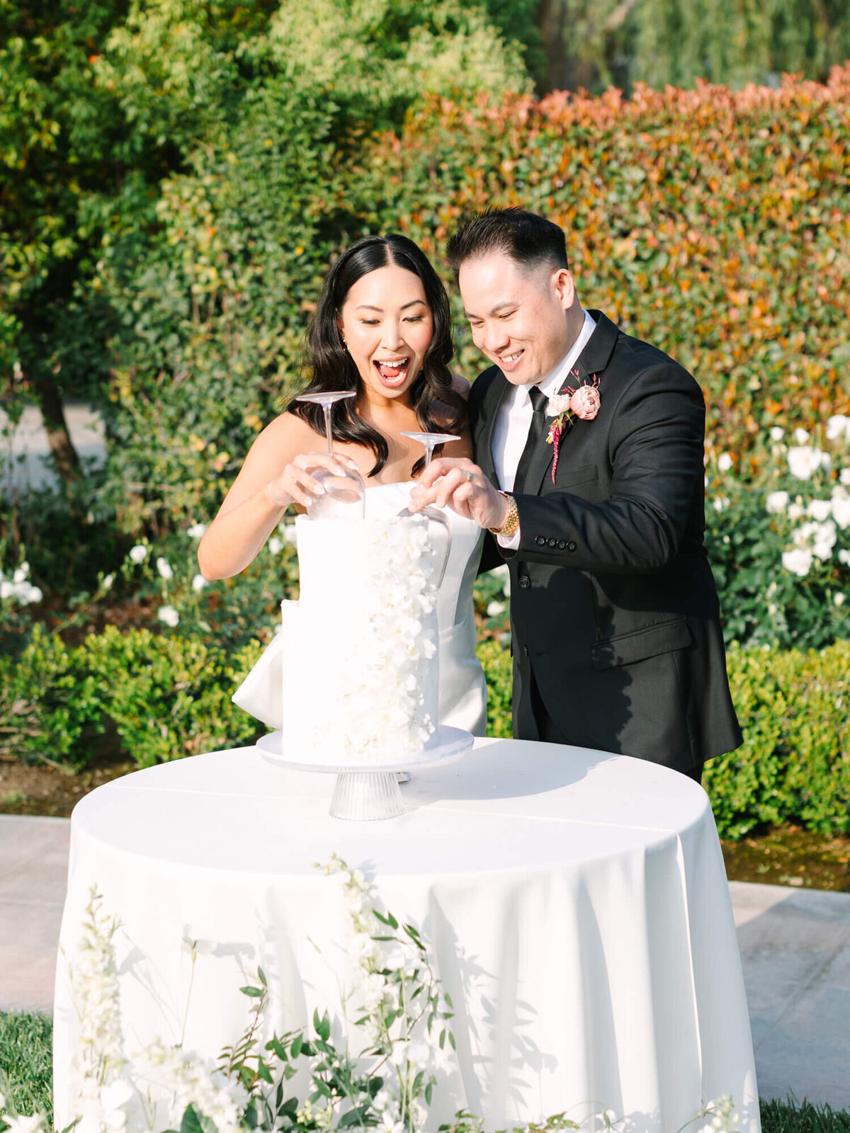 A joyful couple cuts a white floral wedding cake outdoors, surrounded by lush greenery.