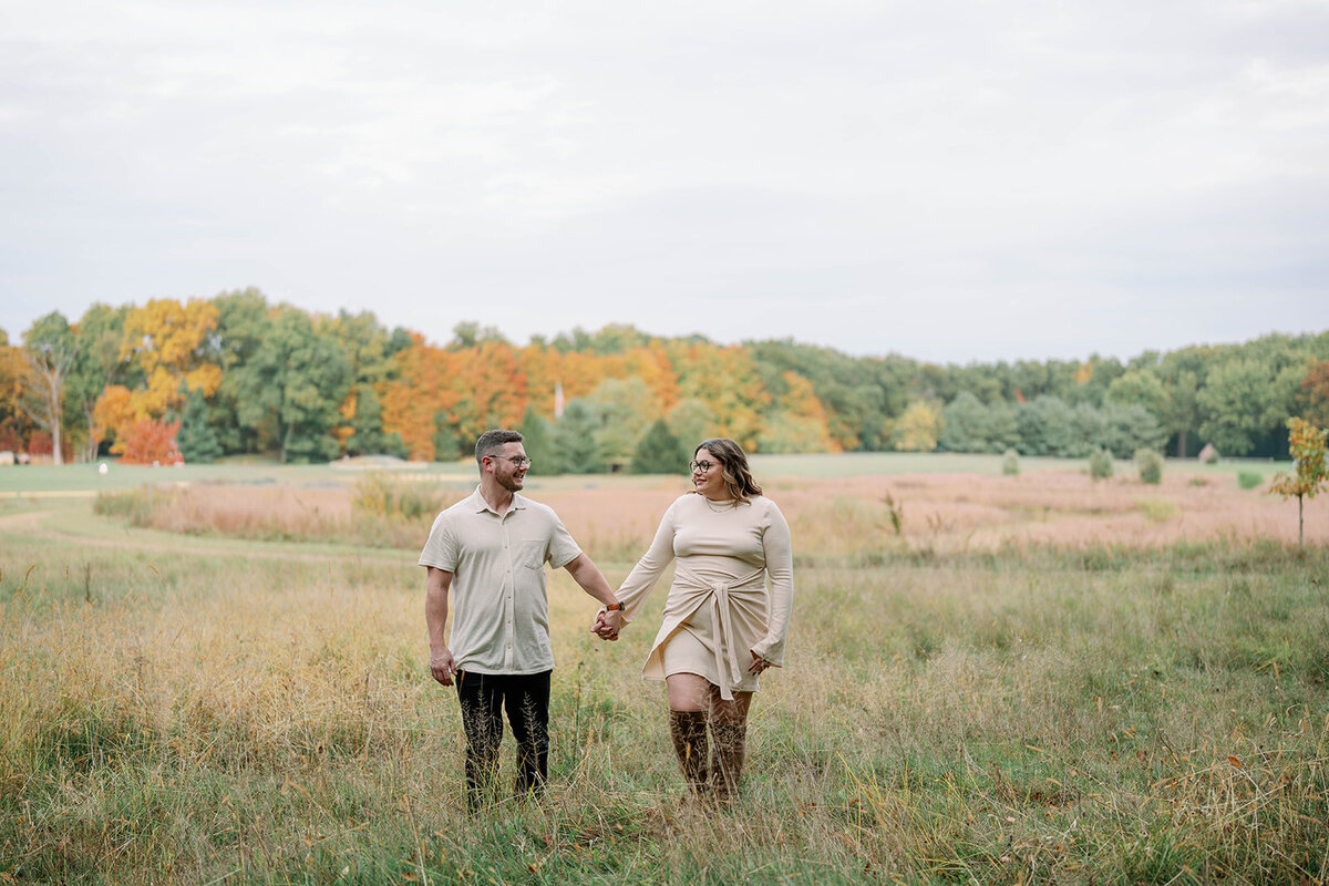 A couple holding hands while walking through an open field with fall foliage during their Michigan engagement session.