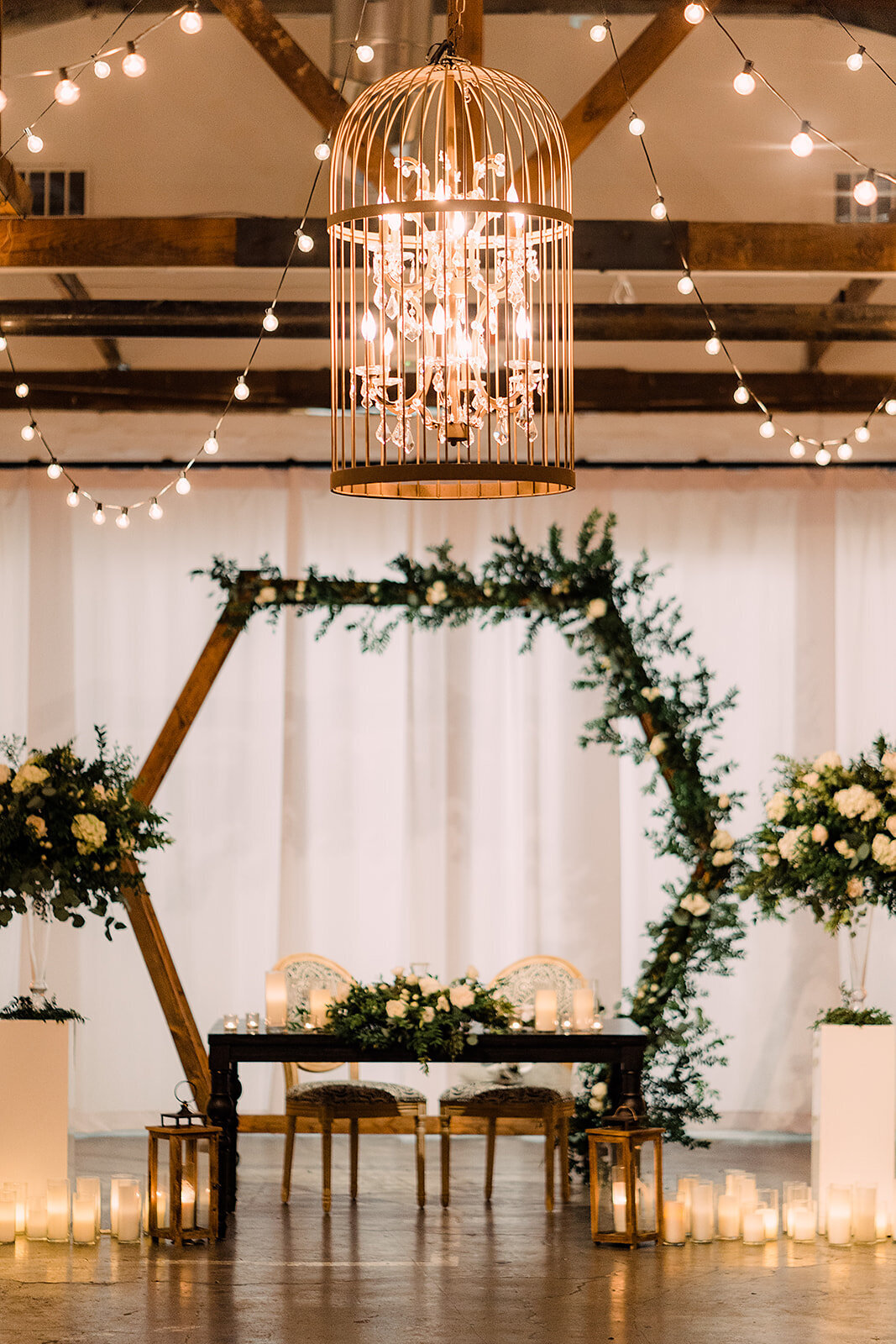 A rustic indoor setting with a wooden wedding arch adorned with greenery and white flowers, framed by string lights and tall pillar candles surrounding a sweetheart table.
