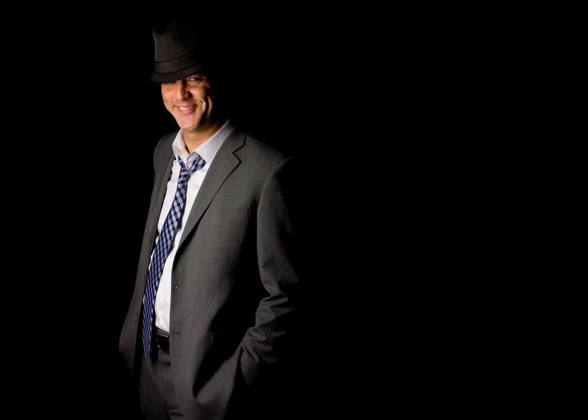 A male dating portrait client in the studio wearing a top hat, smiling & wearing a grey suit