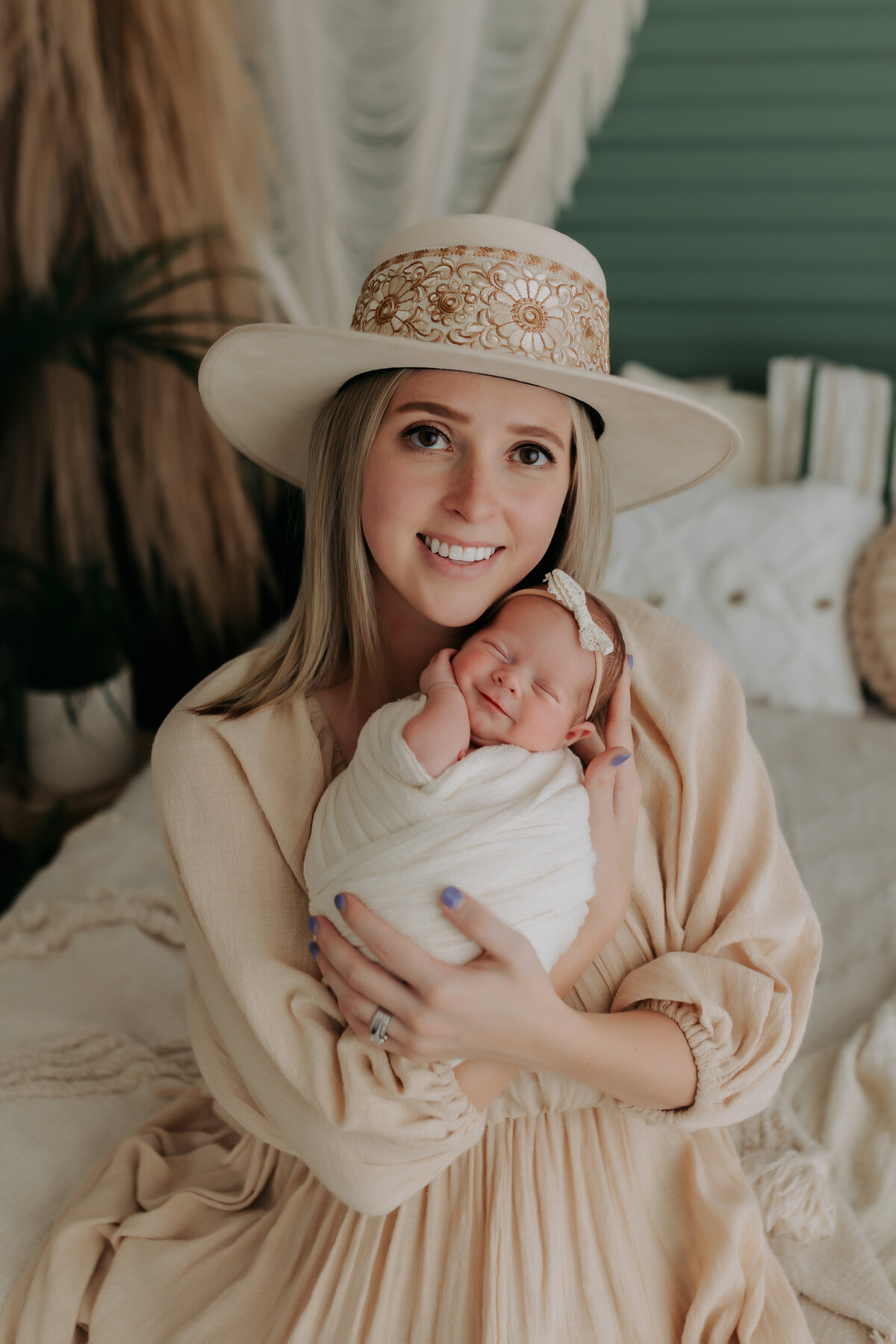 mother wearing bohemian hat holding newborn daughter wrapped in white with a bow