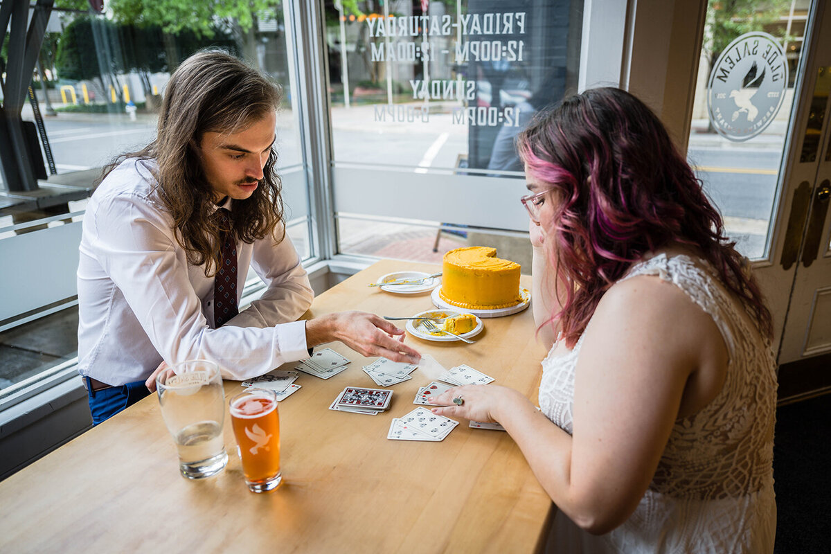 A newlywed couple on their elopement day play a card game together while drinking beers at Olde Salem Brewery and eating vibrant yellow cake.