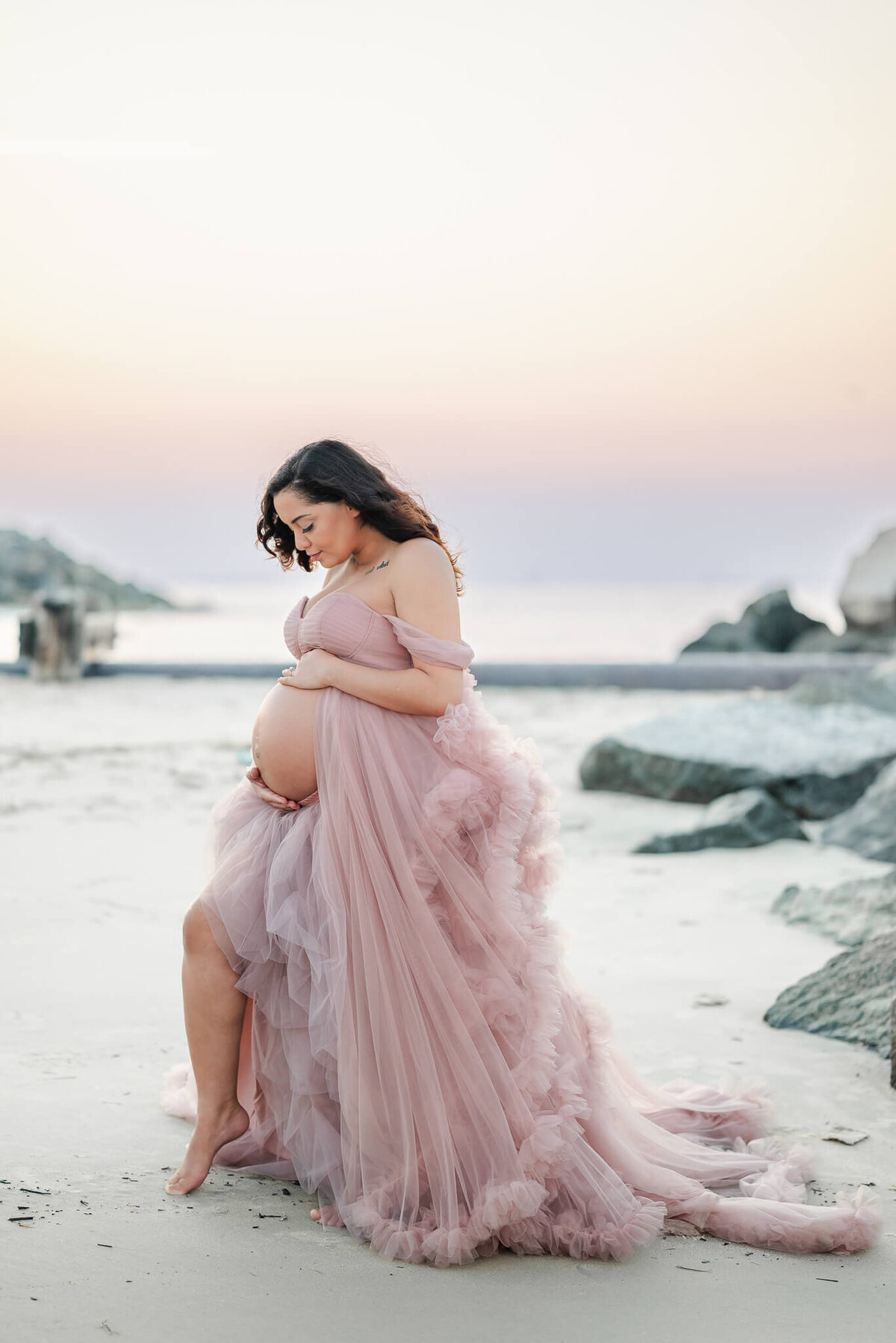 A mam to be is wearing a pink tulle gown during her maternity session. She stands on the beach and looks down at her growing belly.