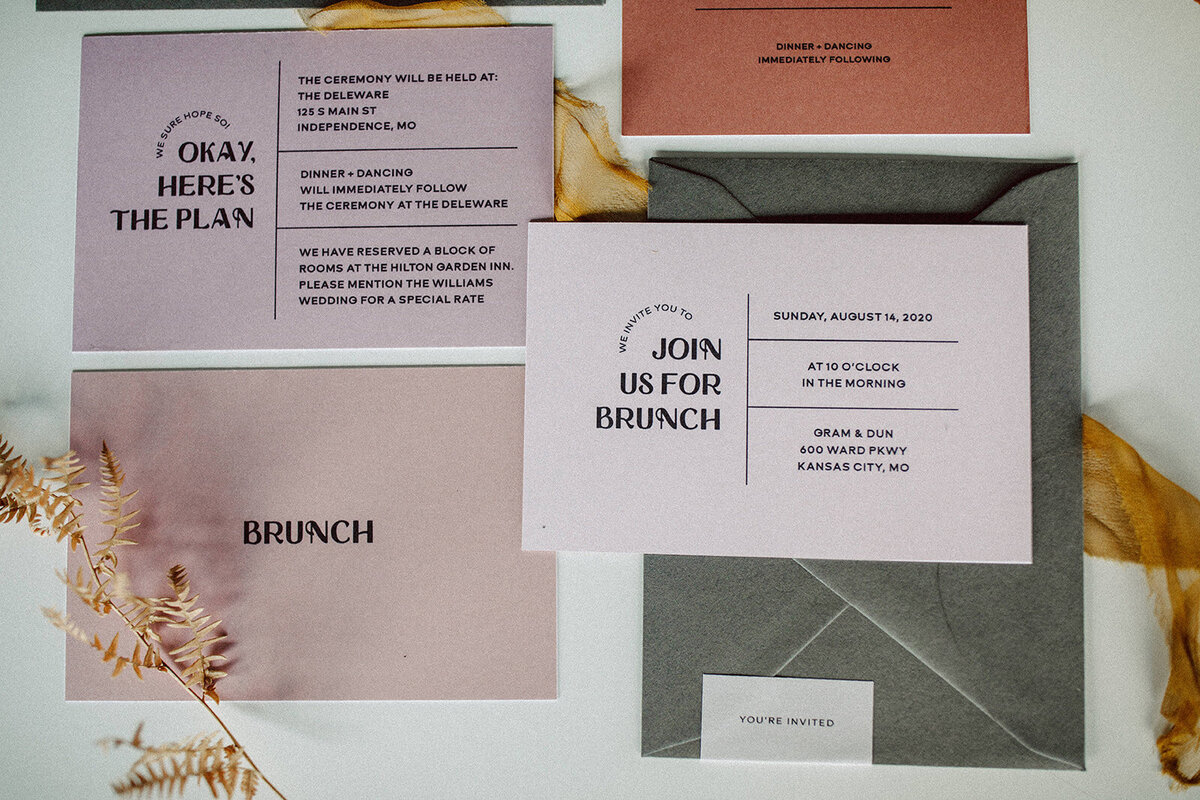 Peach, mauve, and off-white wedding stationery with black font atop a white table with stems and yellow ribbon.