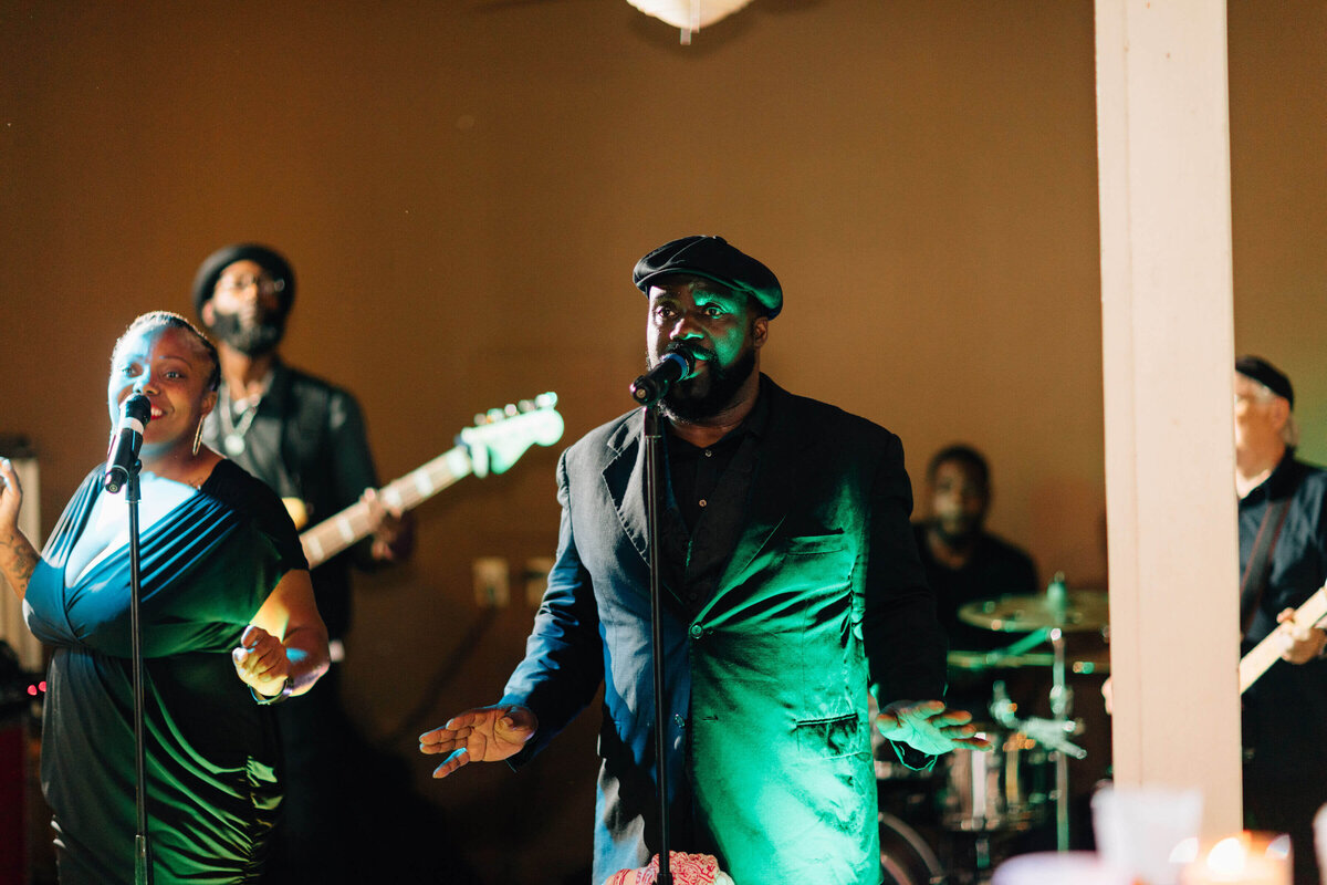 A band performs outdoors. A man in a dark jacket and hat sings into a microphone at center and musicians play guitar and drums in the background.