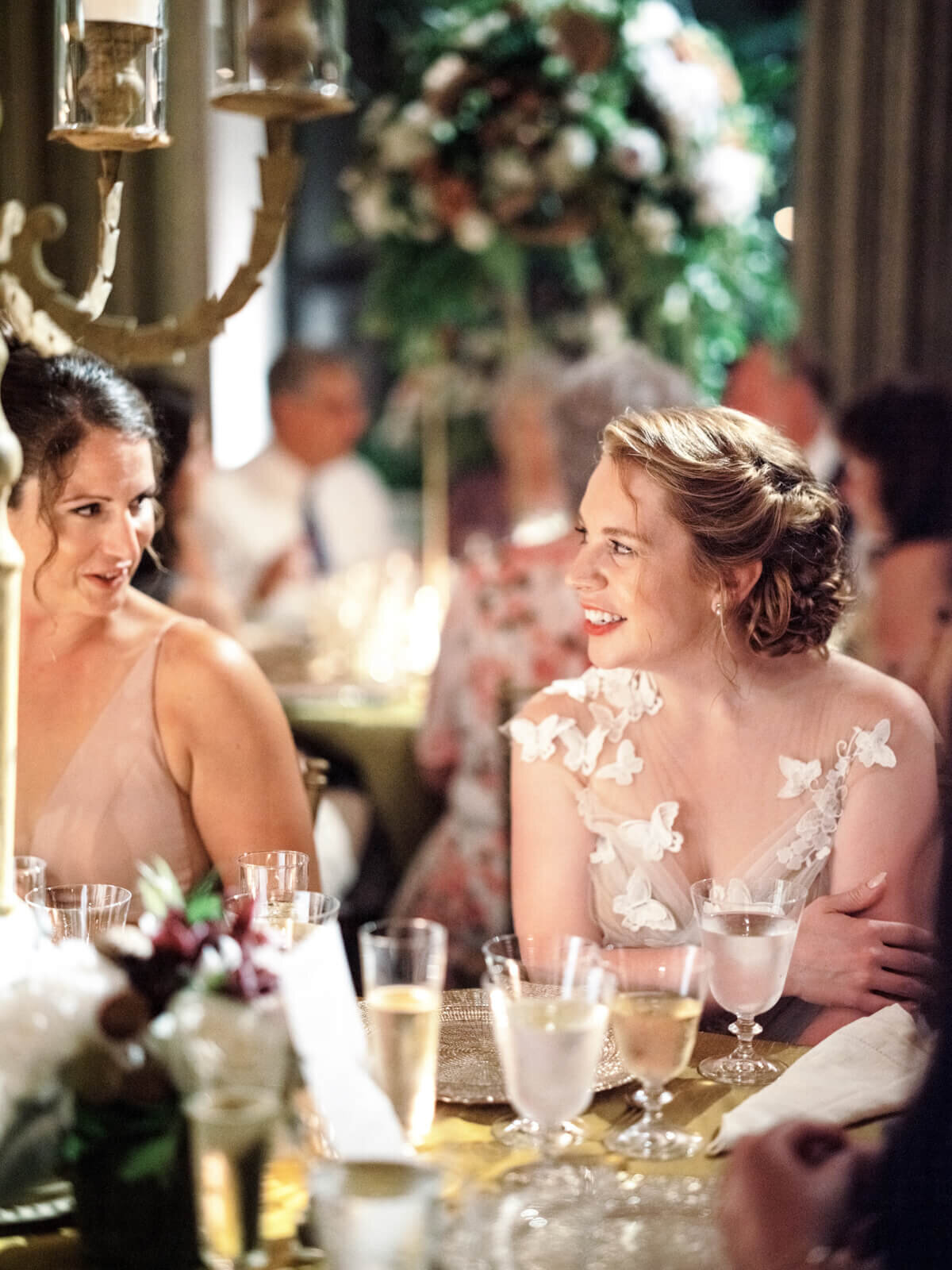 The bride is smiling, looking to the right, seated on a dinner table with a woman beside her; guests are in the background