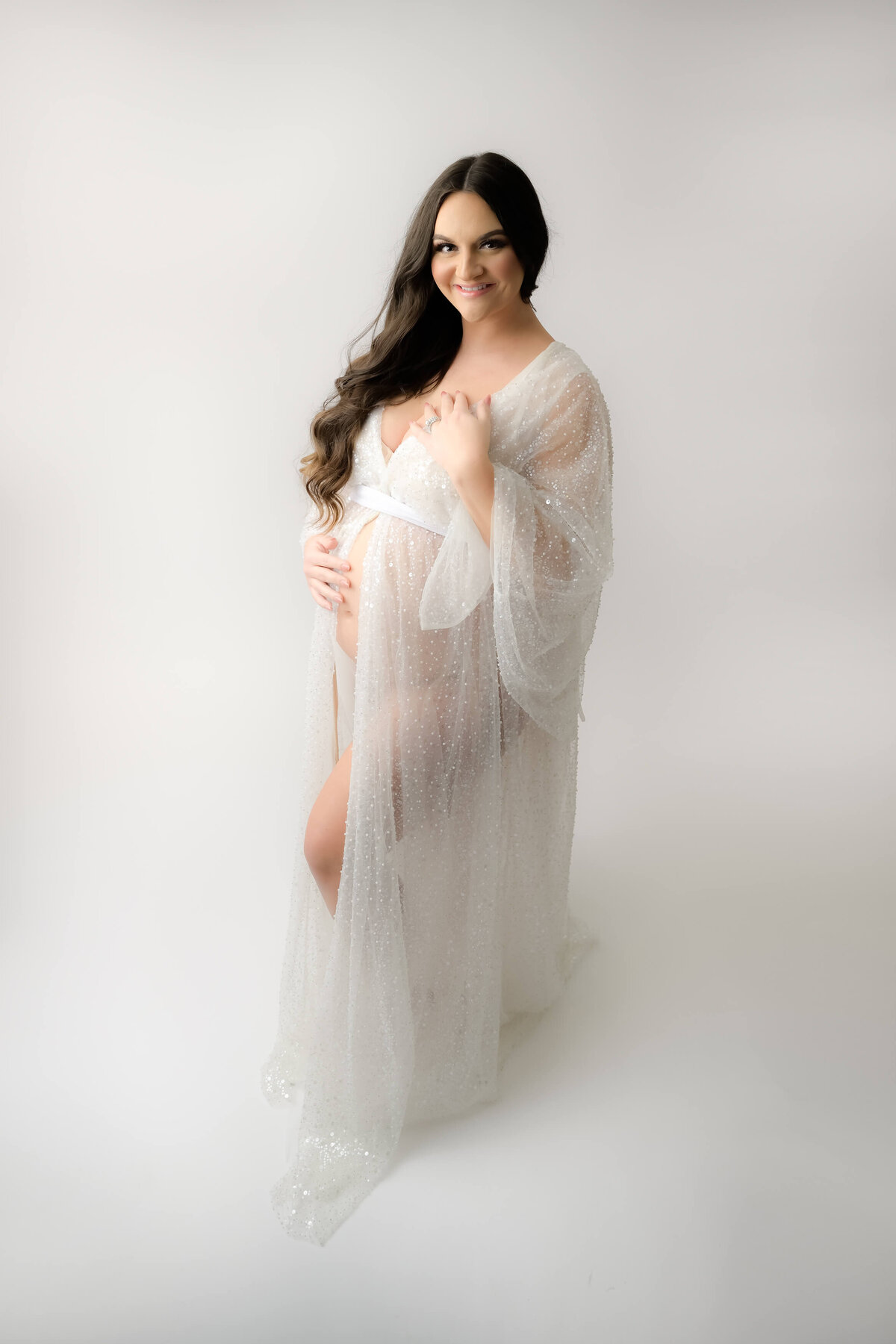 Studio maternity photo session on a white background with expecting mother wearing a stunning white robe with pearls and sequins. Photo taken from slight angle above. Image captured by Beth Benecki