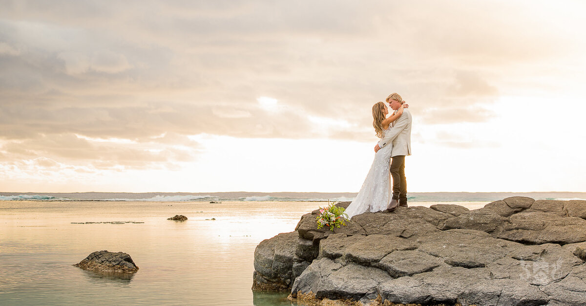 Couple embrace on a rock y foreshore at sunset Rarotonga