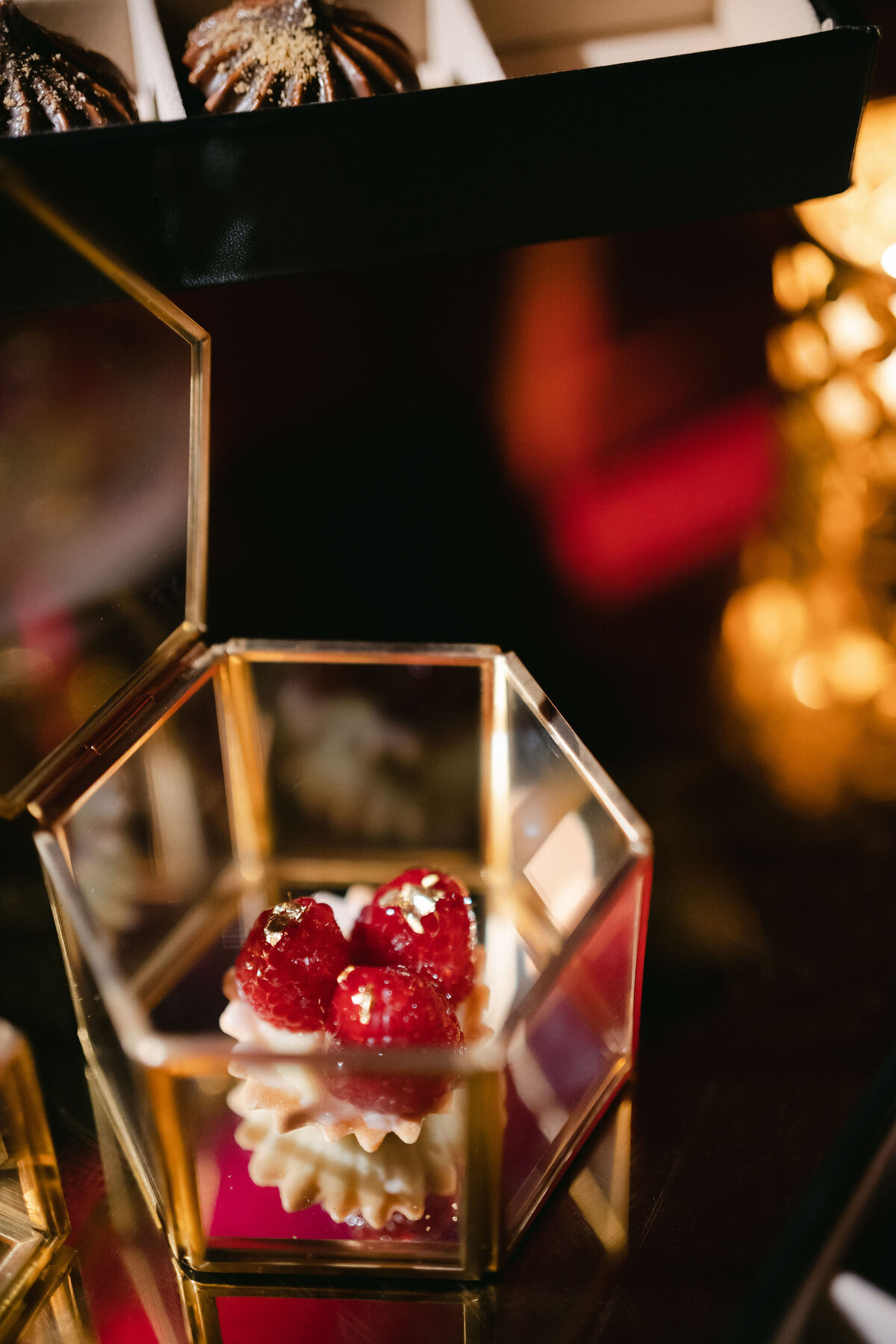 The photo was taken in Virginia, at a tented wedding captured by photographer Elisabetta Marzetti. Clayton Films were present as videographers. Within the frame is a close-up of a delectable dessert displayed in a geometric glass container. The dessert features a delicate pastry adorned with white cream and fresh raspberries, elegantly finished with shimmering gold flakes. In the background, you'll notice blurred elements adding a touch of depth, alongside another similar dessert presented in a box above.