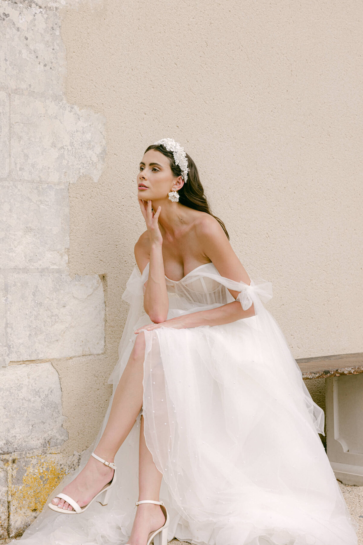 A photo taken during the brides bridal portrait session during her wedding day. She is sitting on a stone bench legs crossed and her chin gently resting on her finger tips she has a beautiful white wedding dress on with white heels and a white floral hair band