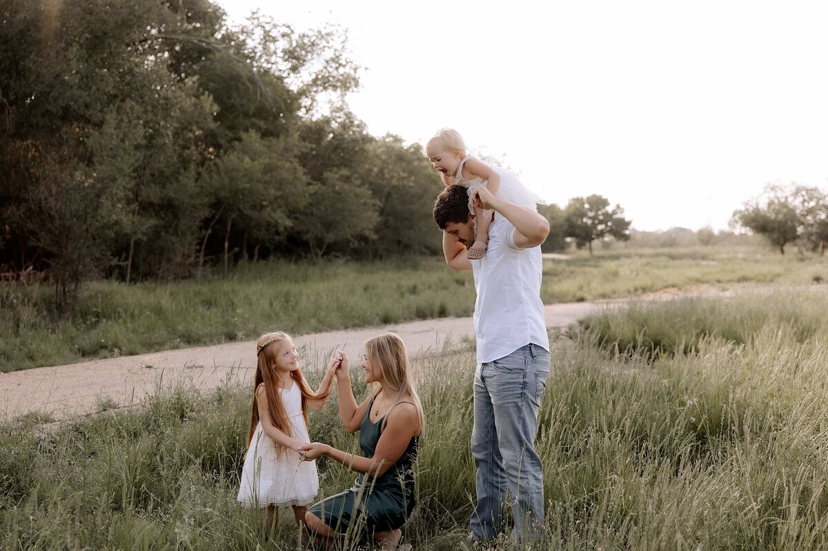 dad has the youngest daughter on his shoulders. Mom is knelt down in the grass playing with the older daughter.