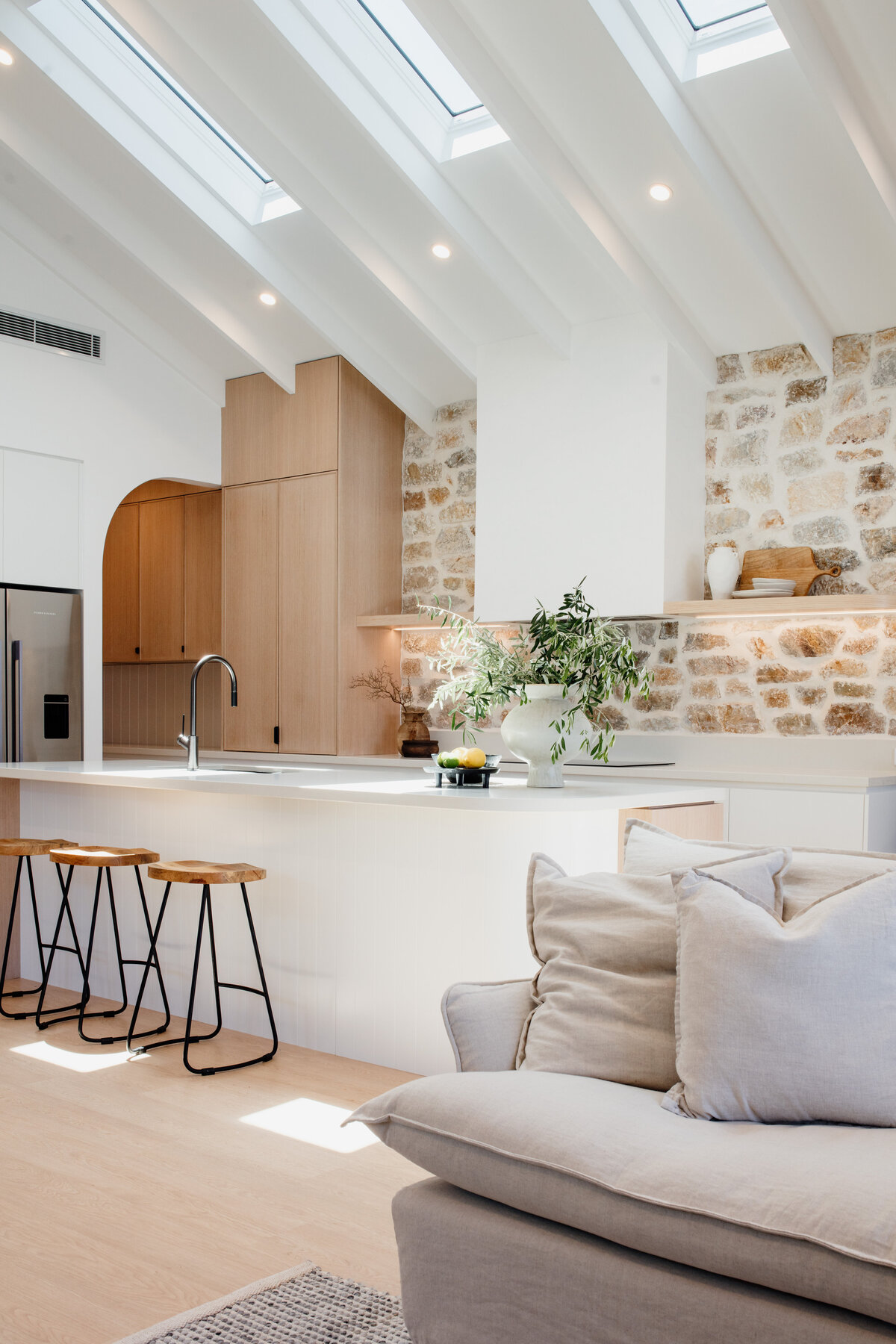 Modern kitchen designed by a renowned Newcastle Interior Designer, featuring wooden cabinets, a stone accent wall, large island with barstools, and skylights. A cozy sofa in the foreground completes the inviting space.