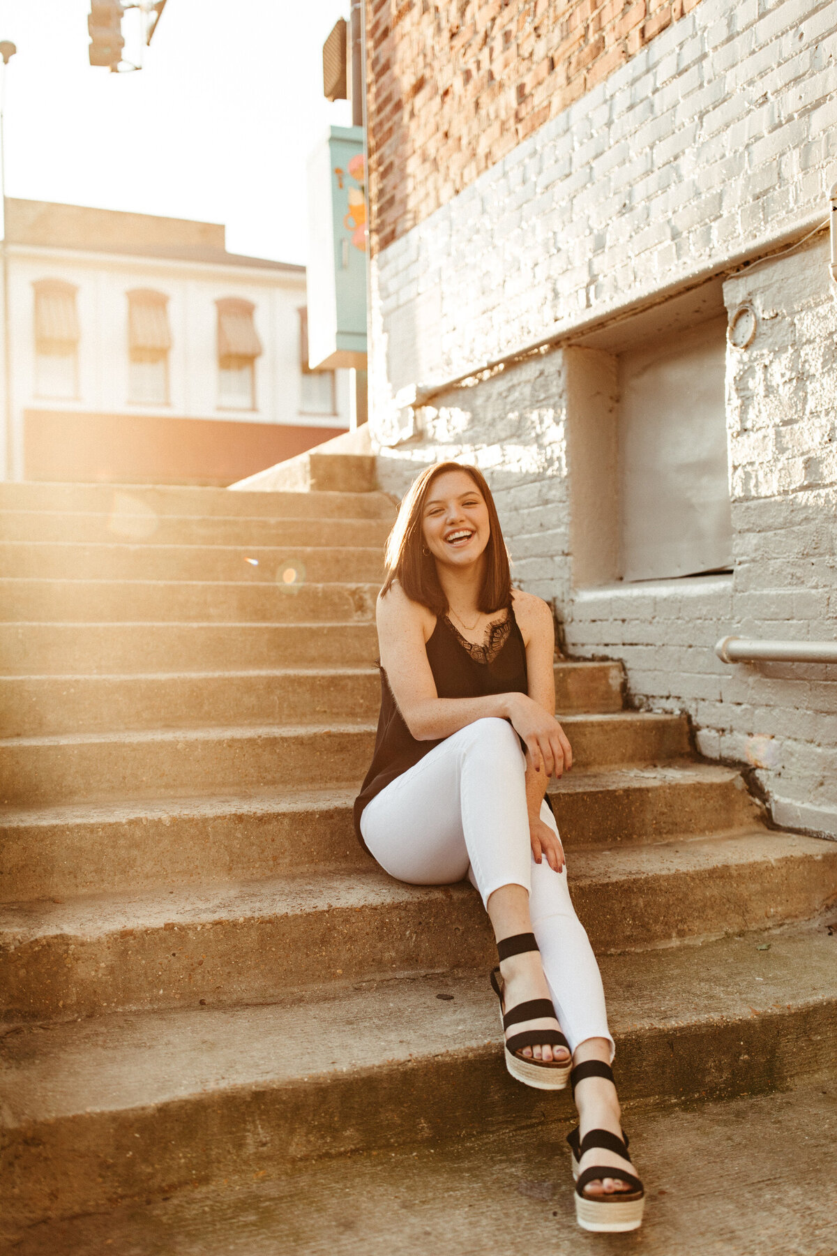 High school senior in white pants and black shirt sitting on stairs downtown at sunrise