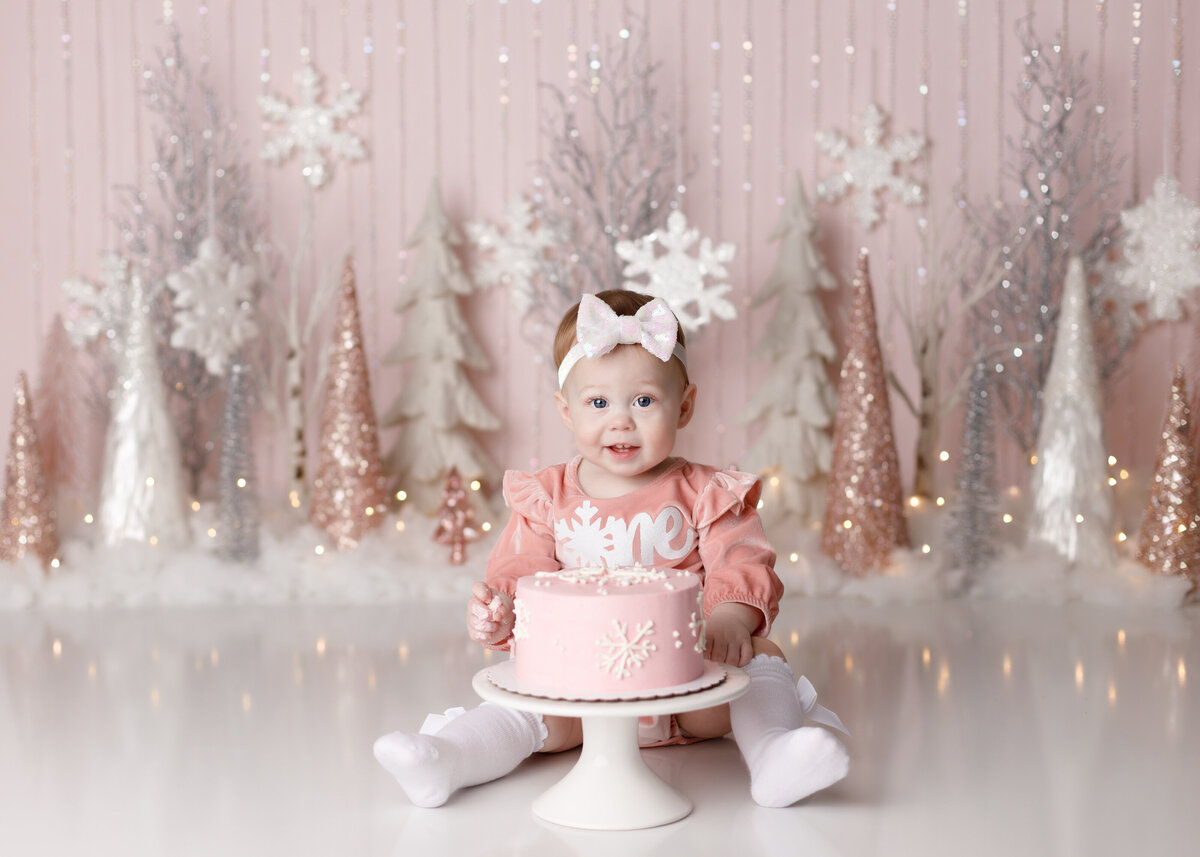 Winter wonderland cake smash in West Palm Beach and Wellington Florida photography studio. Baby girl is wearing a pink onesie with white stockings sitting behind a pink snowflake decorated cake. The background is a soft pink with silver hanging beads surrounded by Christmas tree inspired cones and snowflakes.