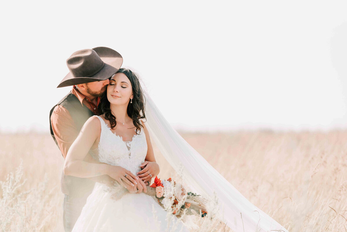 Groom hugs bride from behind and kisses her. 