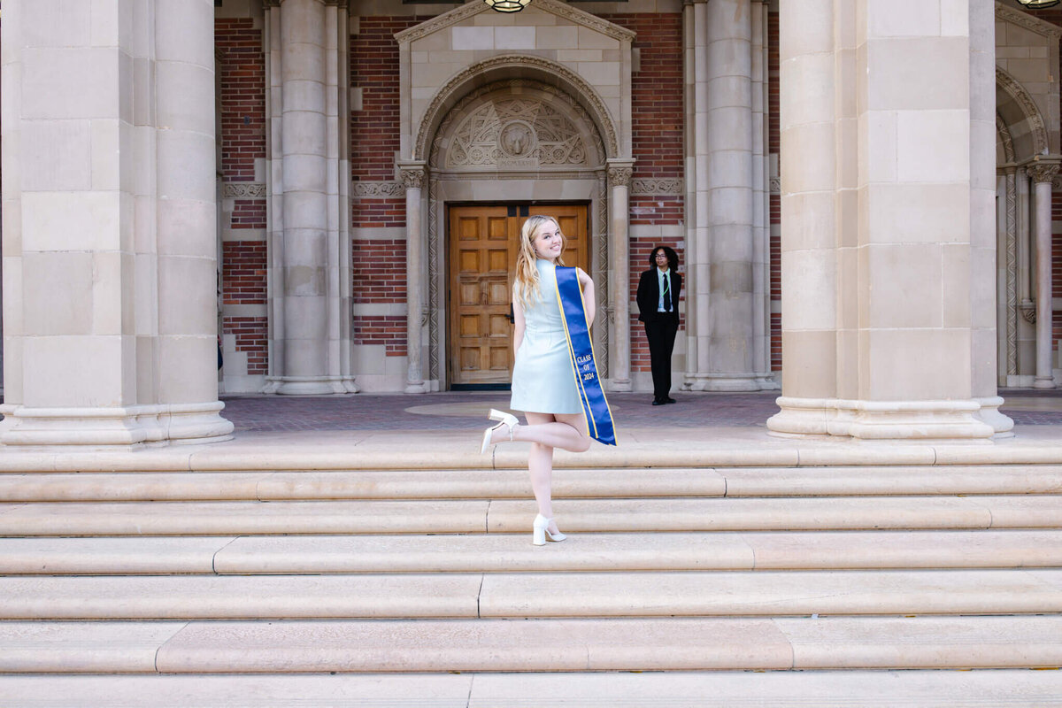 A graduate in a light blue dress and blue sash poses on stone steps in front of a building with large columns. Another person stands in the background near a wooden door.