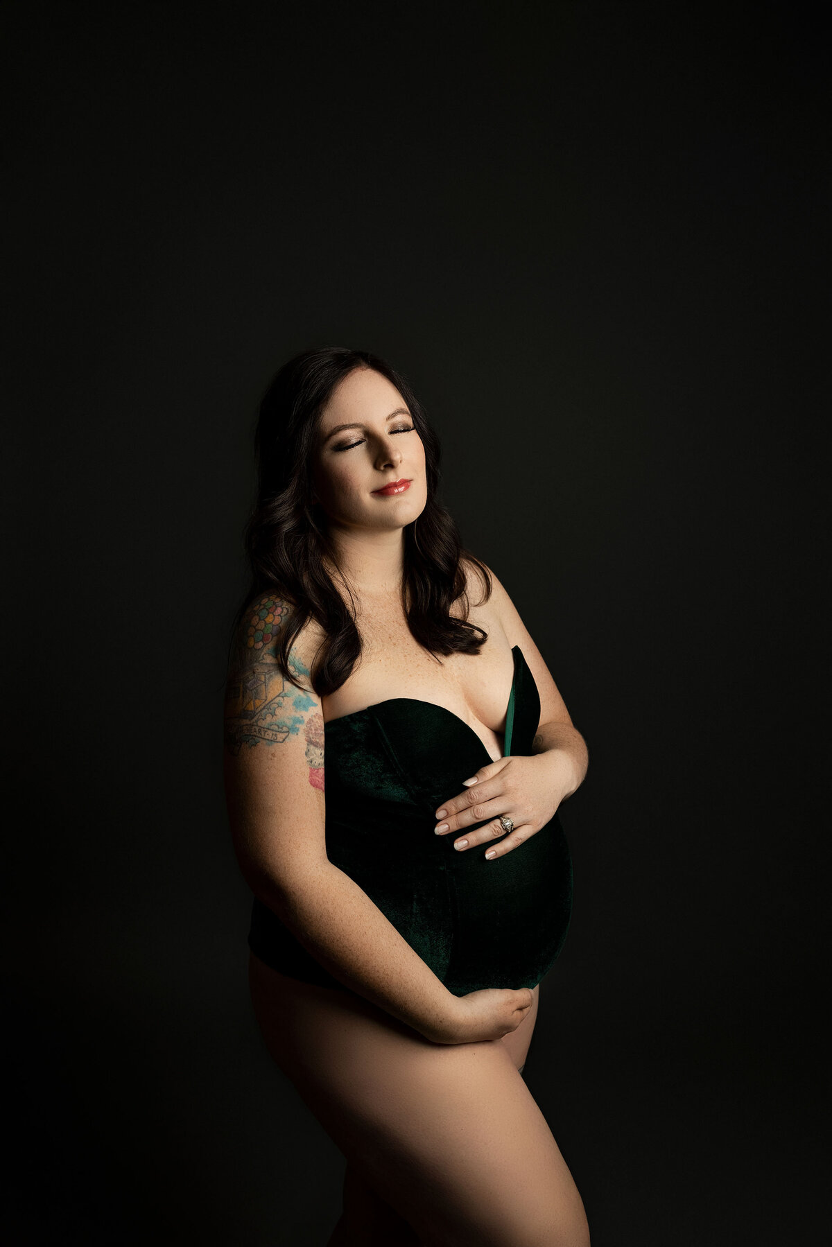 Expectant mom in deep green velvet bodysuit is holding her baby bump in london, ontario materntiy photoshoot. Mom's eyes are closed and her face is angled up to the light. She has bare legs. She is standing against a dark greyish black background. Her forearm has colourful tattoos.