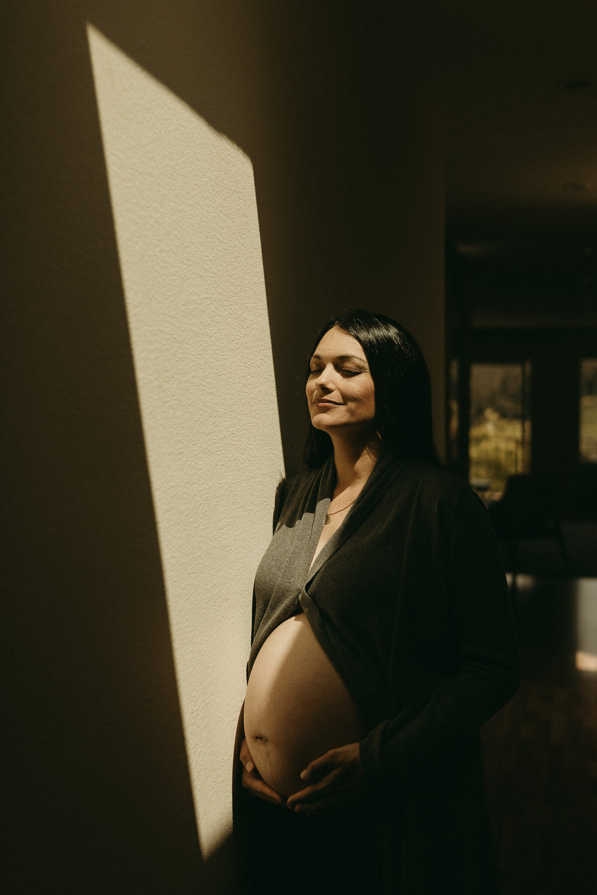 Serene maternity capture with woman holding bare belly eyes closed and triangle of light on wall