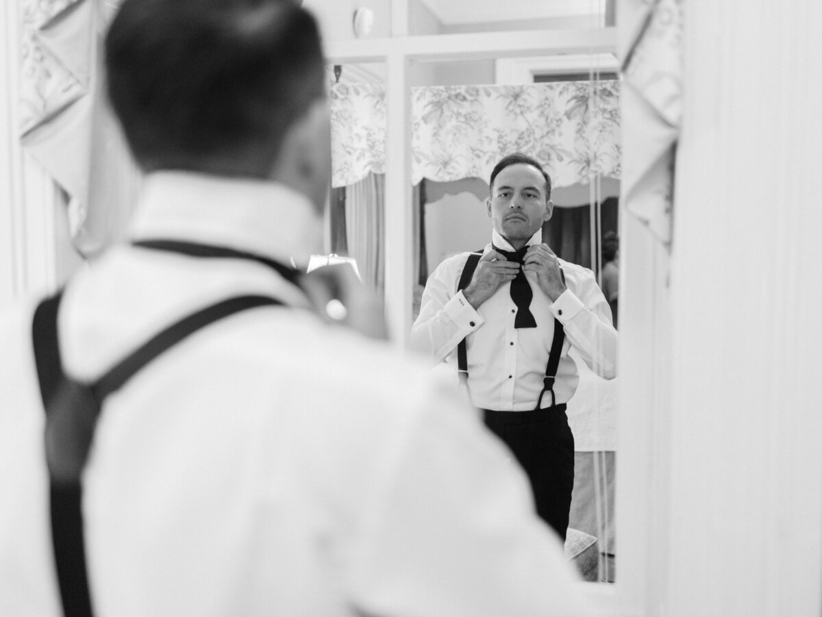 The groom confidently adjusting his bowtie in front of the mirror, representing determination and attention to detail as he prepares for his important day ahead.