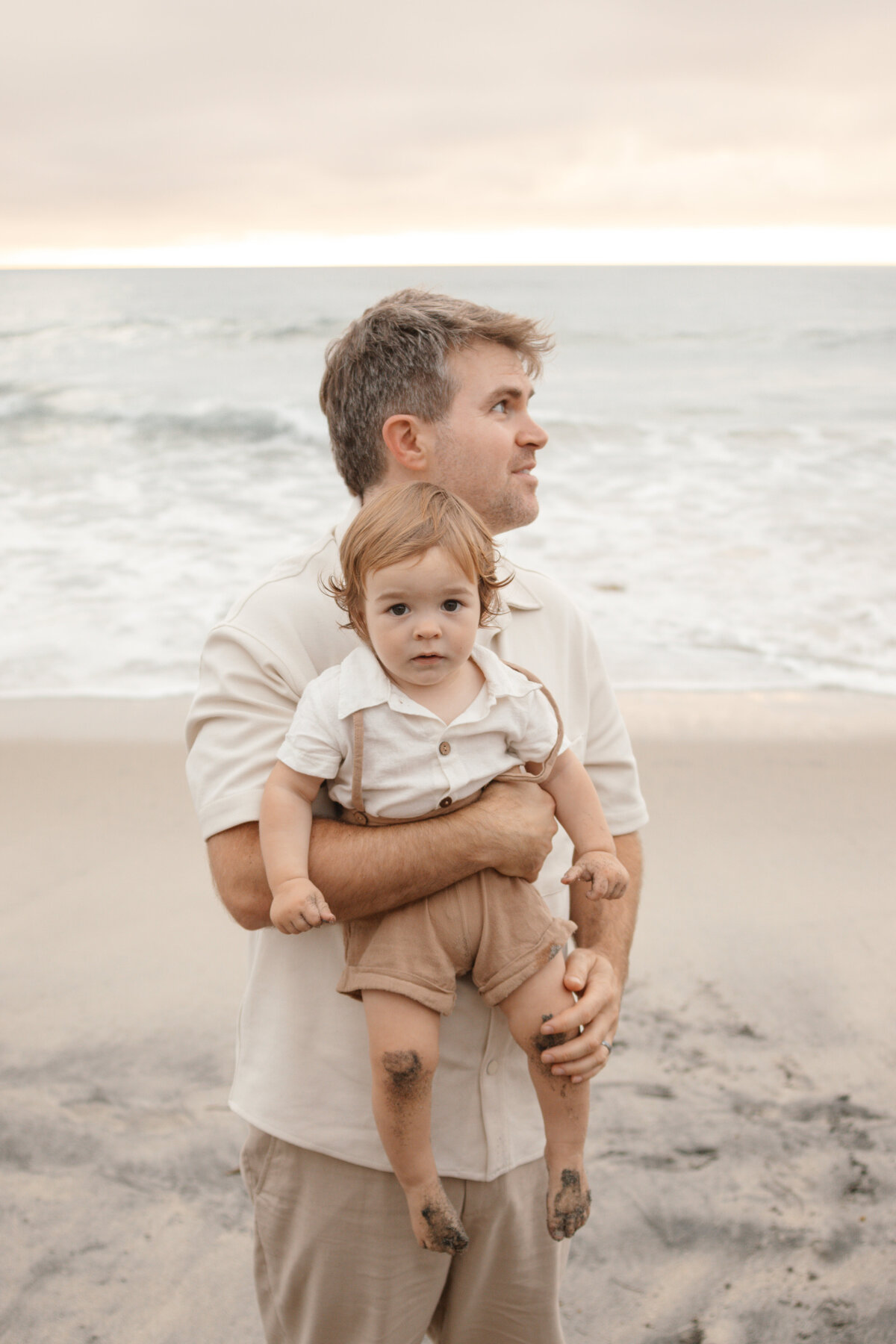 dad holds son who has sandy knees and toes from the beach while dad looks off into the distance
