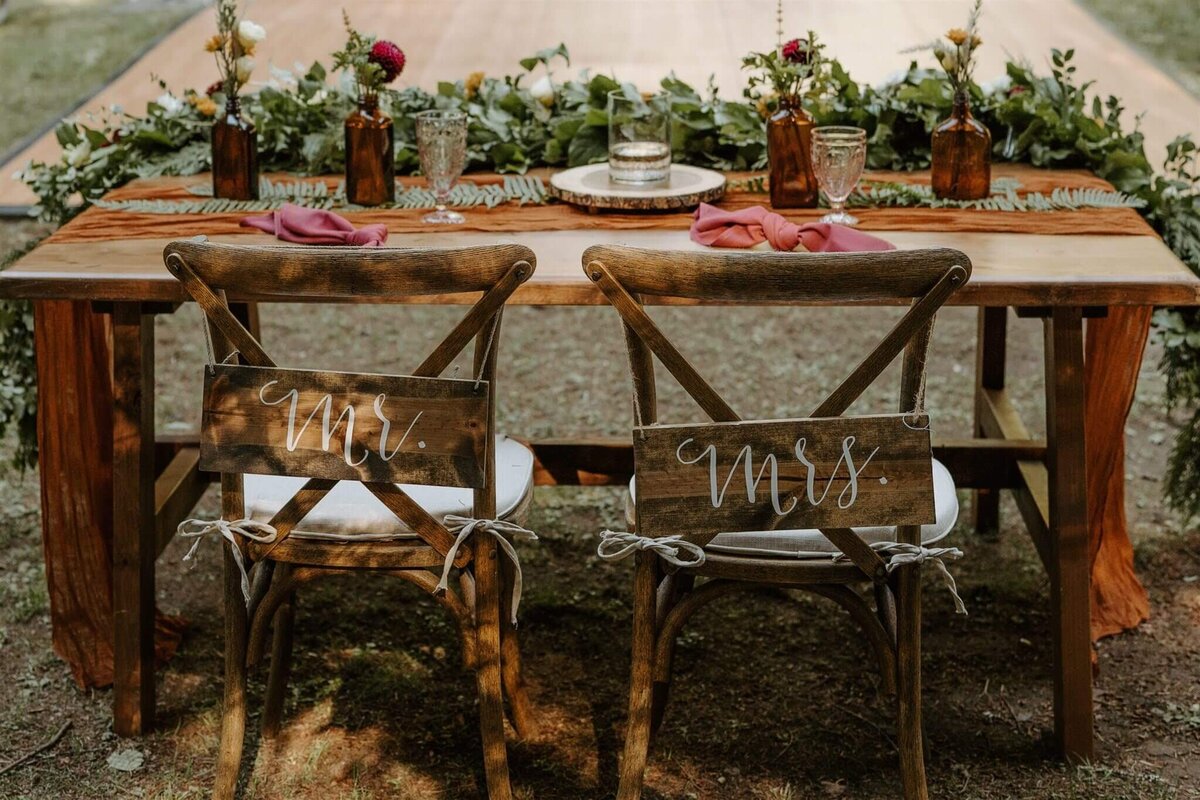 The sweetheart table at a wedding reception is shown with bud vase arrangements and a floral garland and mr and mrs signs.