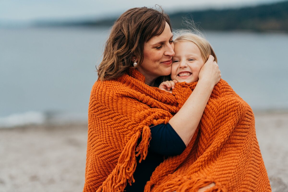 mom-snuggling-with-daughter-under-a-blanket-near-beach