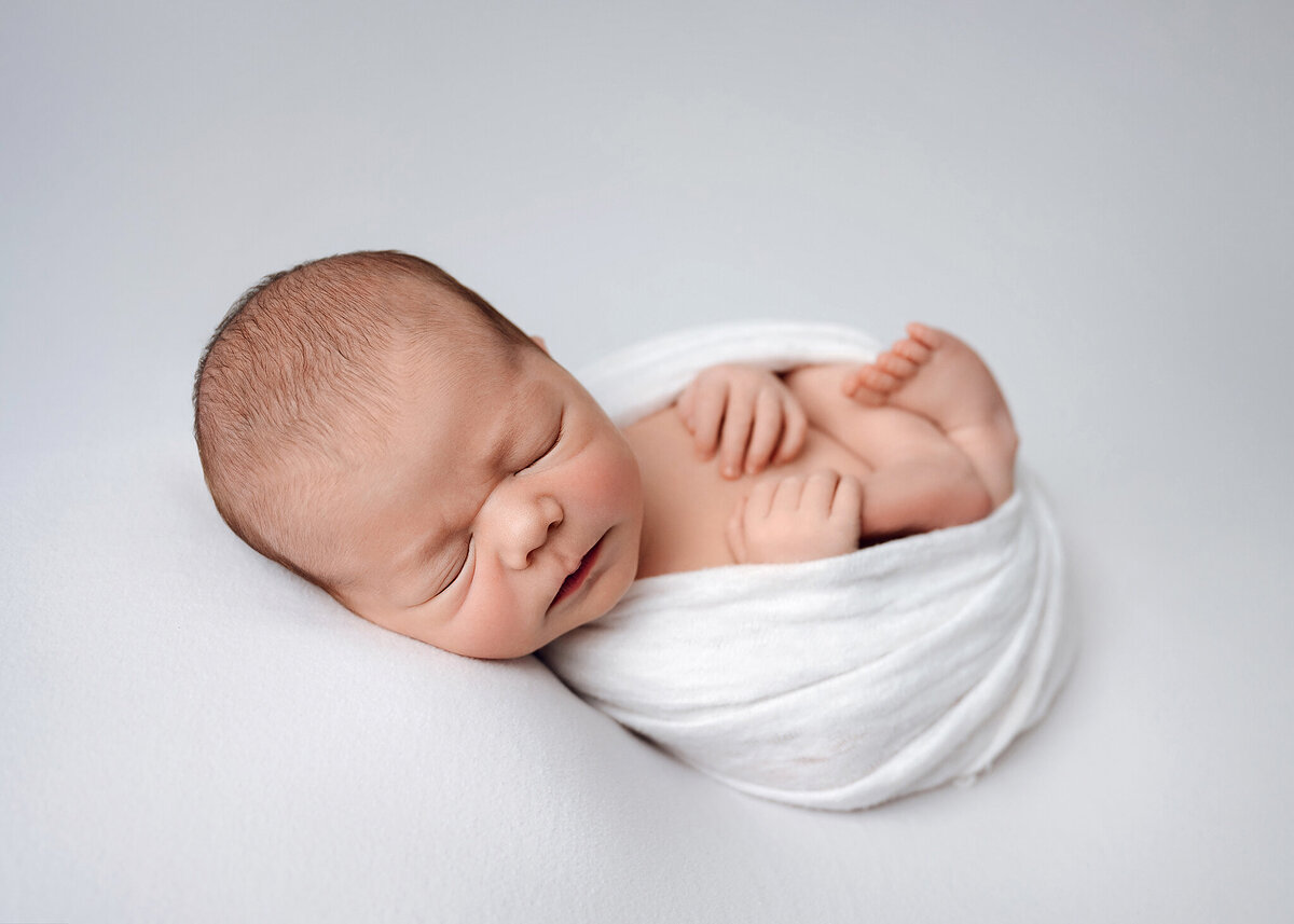 baby boy swaddled with his feet and hands out  sleeping on a white blanket in a simple studio set up