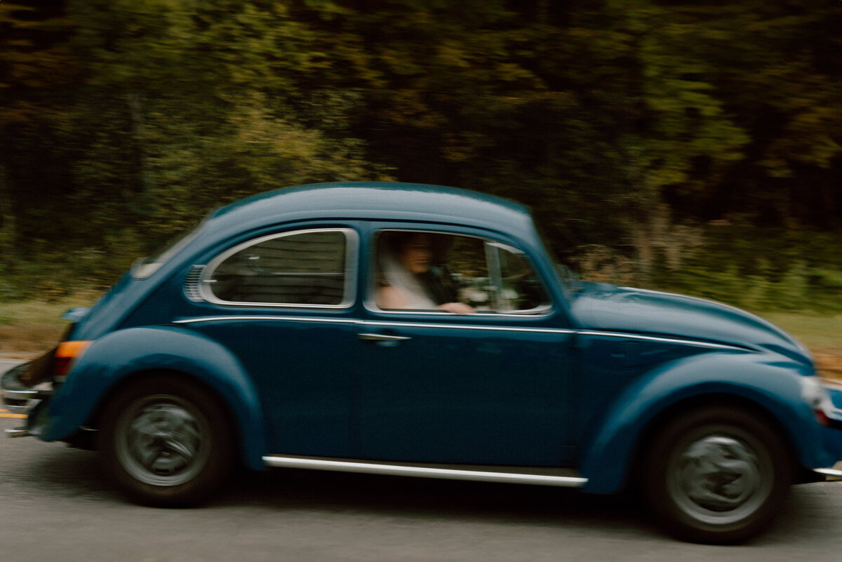 Bride and groom captured in motion as she drives a classic blue Volkswagen Beetle, showcasing a candid, documentary style. The blurred background of lush greenery emphasizes the car's movement, creating a dynamic and nostalgic wedding day moment.