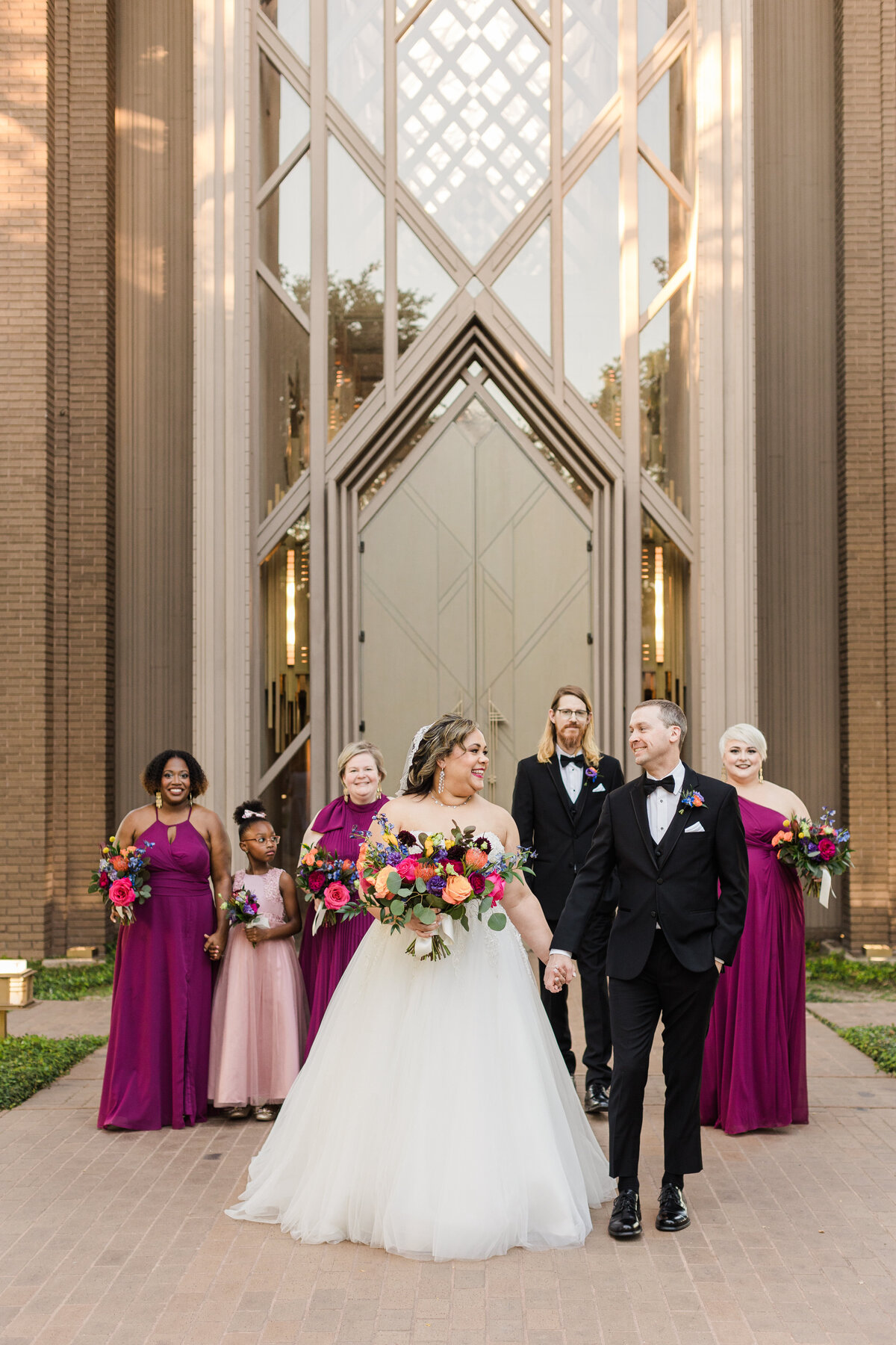Portrait of a bride, groom, and their wedding party posing outside of the beautiful Marty Leonard Community Chapel in Fort Worth, Texas. The bride is on the left and is wearing a flowing, white, sleeveless dress with a long view and is holding a large, colorful bouquet. The groom is on the right and is wearing a black tuxedo with a bowtie and a boutonniere. The women in the wedding party are wedding burgundy or light pink dresses and all have bouquets. The one man in the wedding party is wearing a black tuxedo with a bowtie and boutonniere.