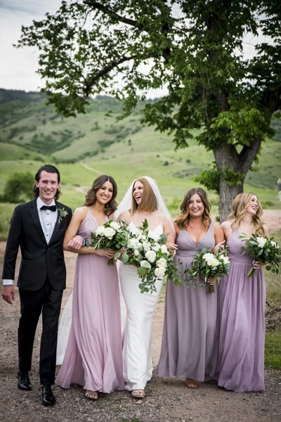 A bride walks arm in arm with her bridal party as she laughs at one of her bridesmaids.