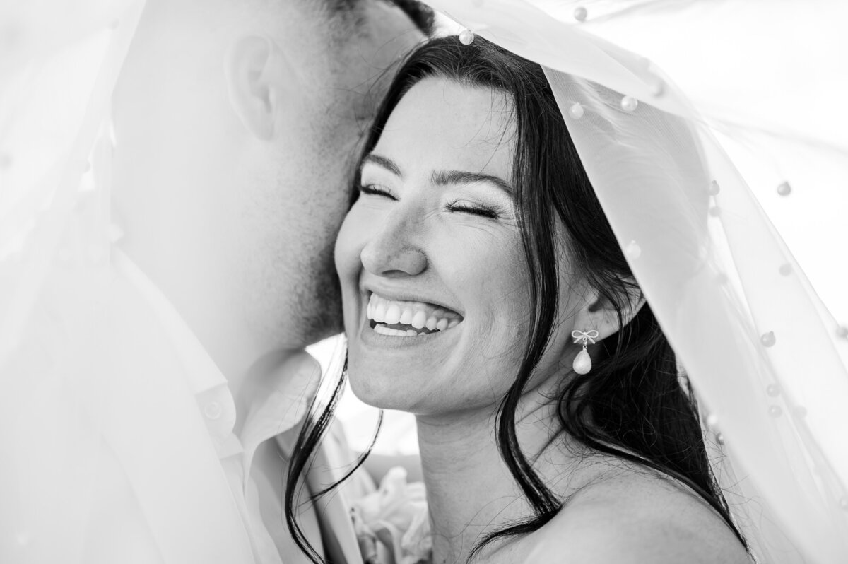 A laughing bride under her wedding veil as her husband whispers in her ear in a black and white photo by Raleigh wedding photographer, JoLynn Photography